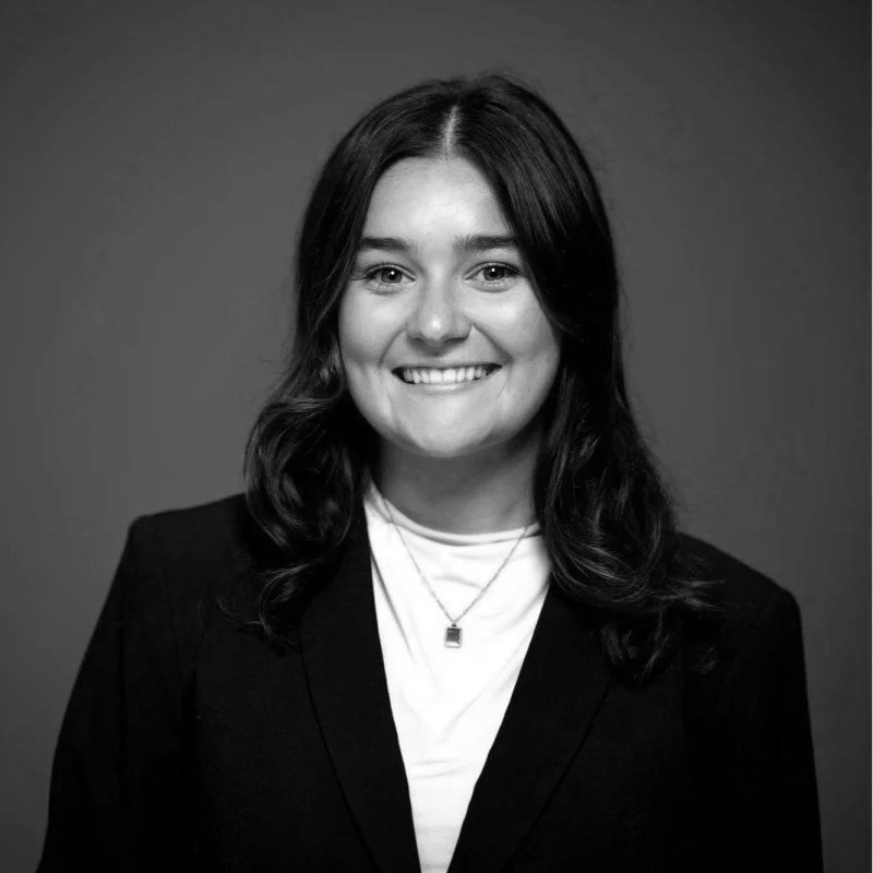 Black and white portrait of a woman smiling, with shoulder-length wavy hair, wearing a blazer over a t-shirt and a necklace, against a plain background.