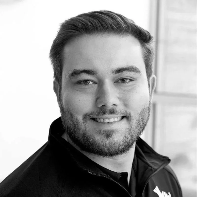 A smiling man with light hair and a beard, wearing a dark jacket, in front of a window.