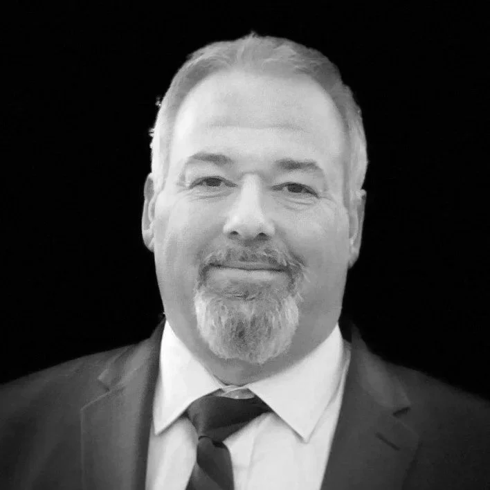 Black and white photo of a smiling middle-aged man with short hair, a beard, dressed in a suit and tie, against a dark background.