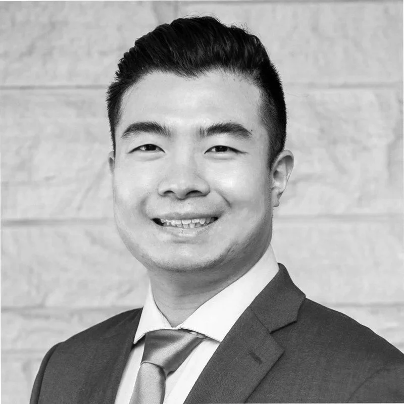 A black-and-white professional portrait of a young man wearing a suit and tie, smiling, with a wooden wall background.