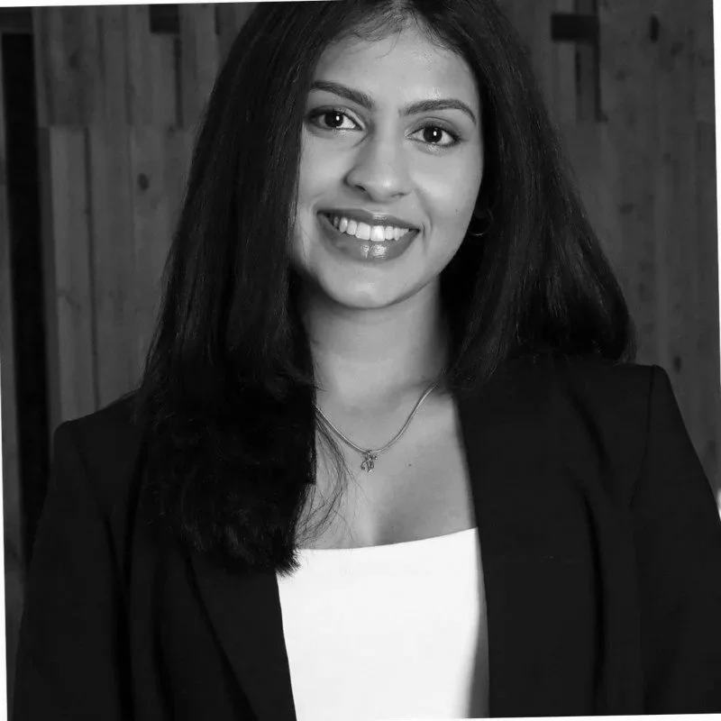 Black and white portrait of a young woman with long dark hair, smiling, wearing a blazer and a necklace, in front of a wooden background.