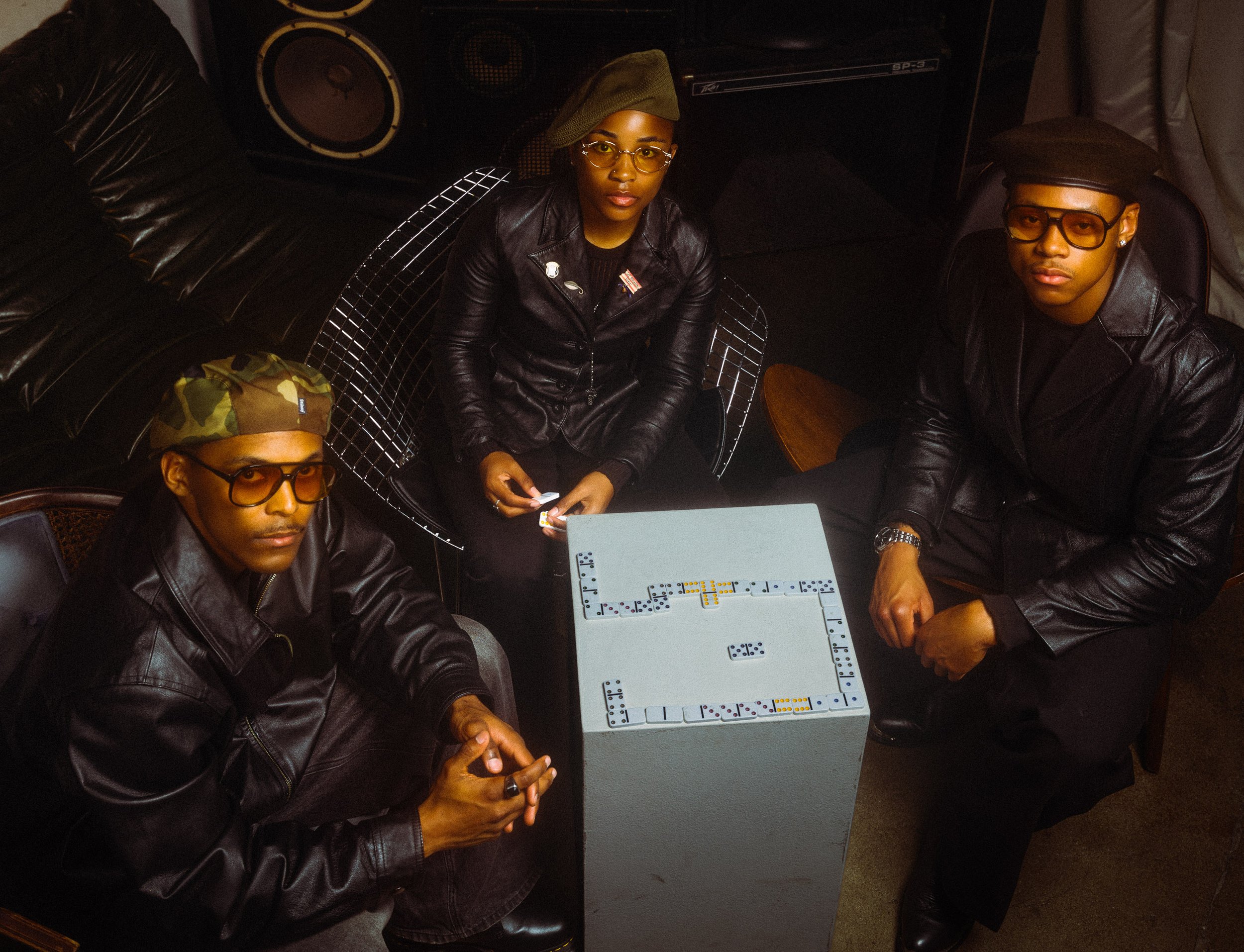 Three people sitting around a table playing dominoes, all wearing black leather jackets and sunglasses, in a dimly lit room.