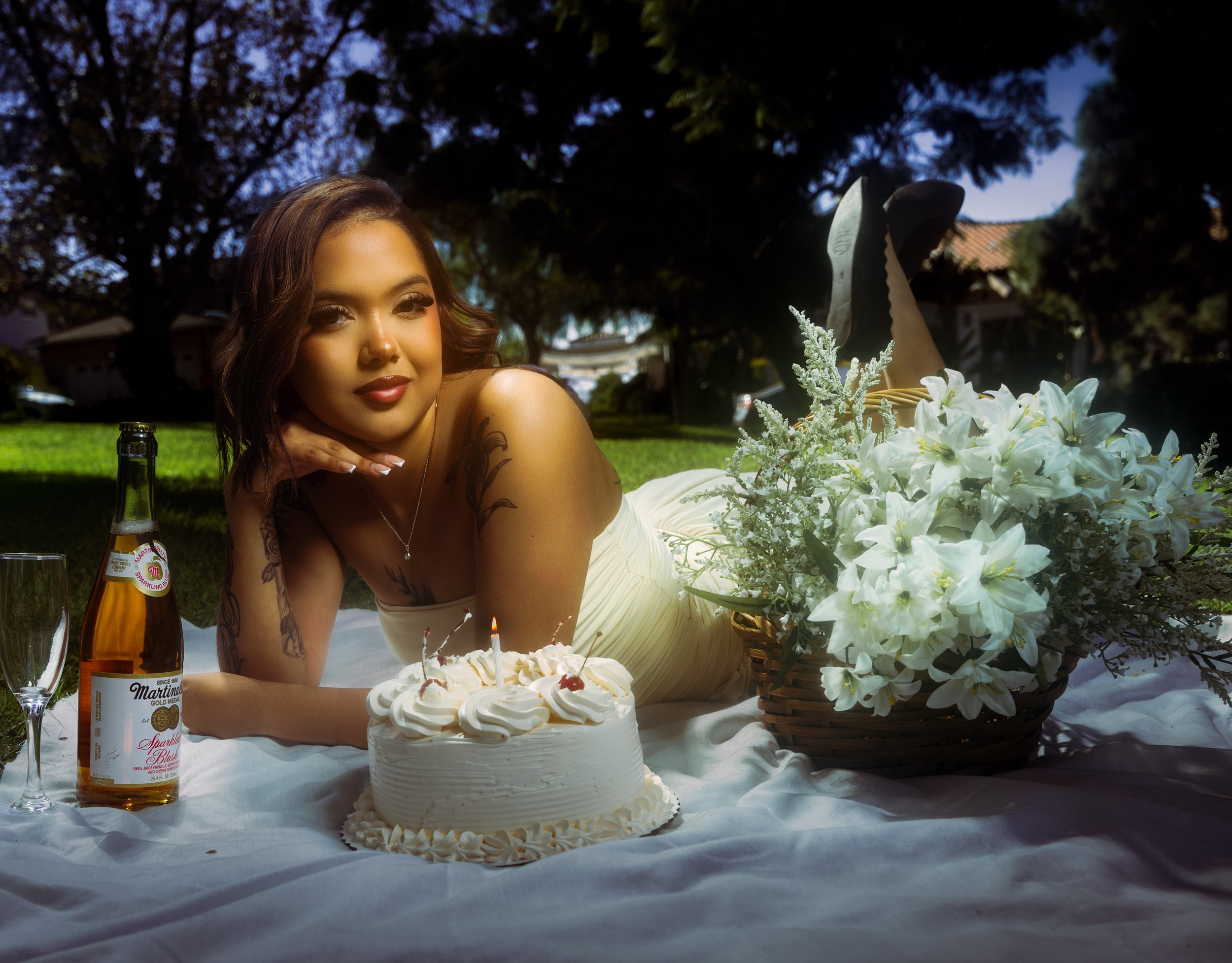 A woman with shoulder-length hair and tattoos lying on her stomach on a white blanket outdoors during the daytime, posing with a birthday cake, a bottle of sparkling beverage, a glass, and a large basket of white lilies and other flowers.