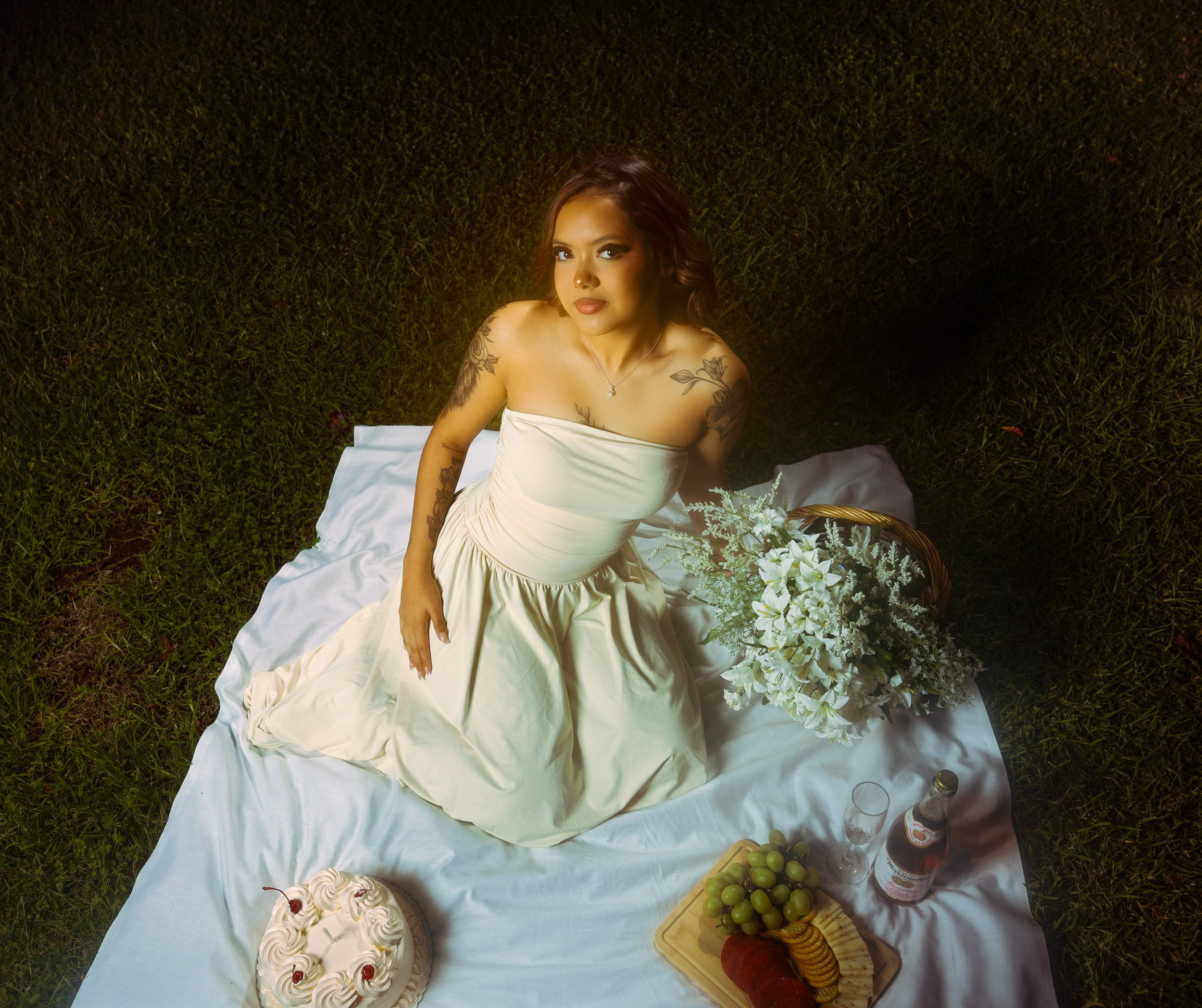 A woman in a strapless white dress is sitting on a white blanket outdoors on grass. She is looking up at the camera with tattoos on her arms and shoulders. Next to her is a bouquet of white flowers, a cake with cherries, a plate of grapes and sliced 