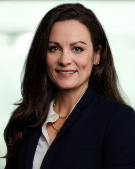 Professional woman with long dark hair wearing a navy blazer and white shirt, smiling at the camera in an office environment.