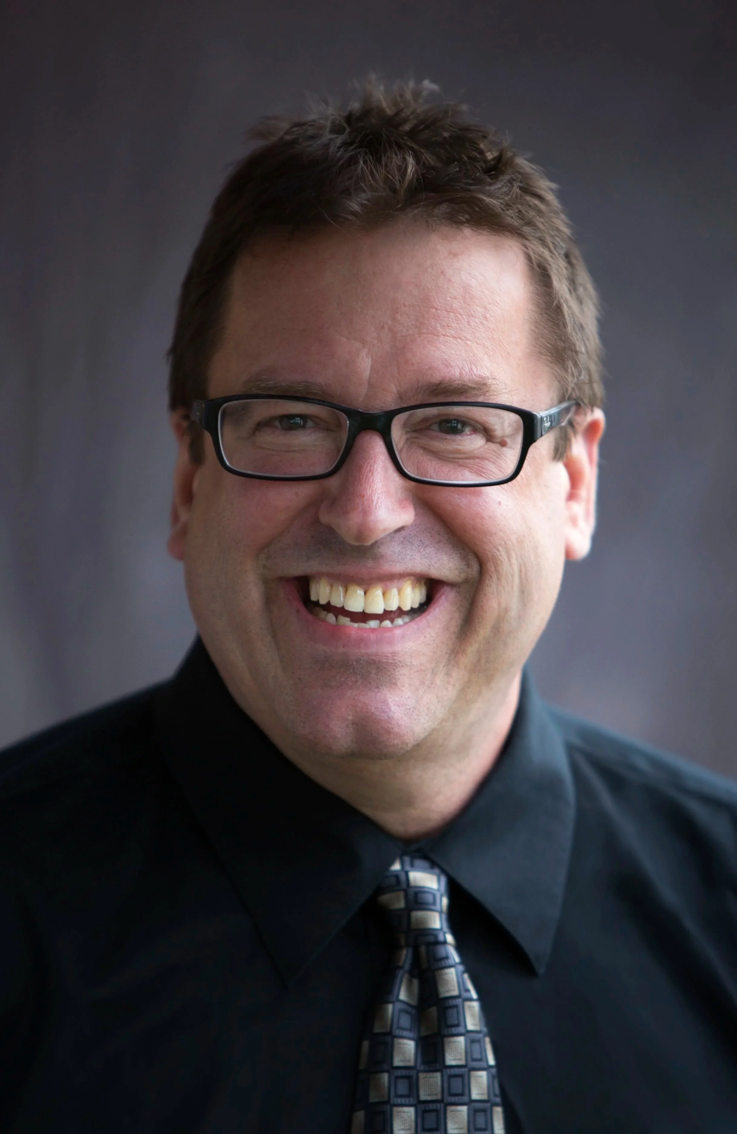 A smiling man with glasses wearing a dark shirt and patterned tie.