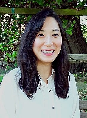 A woman with long black hair wearing a white button-up shirt, standing outdoors with greenery and a wooden fence in the background.