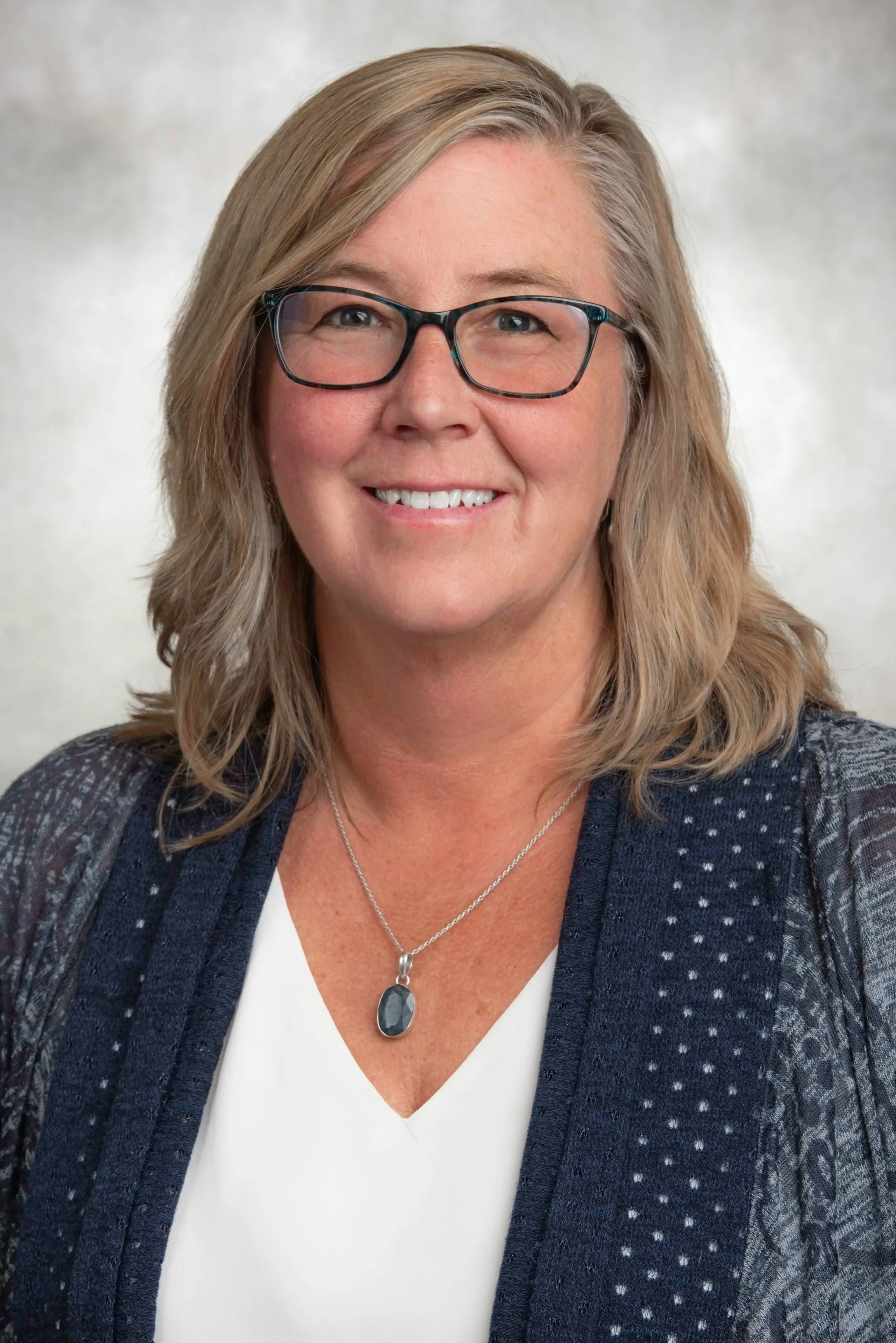 Headshot of a middle-aged woman with shoulder-length blonde hair, glasses, and a friendly smile, wearing a white top, a dark patterned blazer, and a necklace with a dark stone pendant.