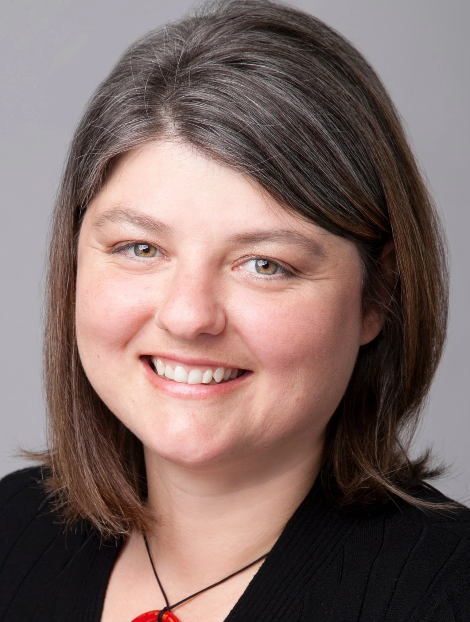 Close-up photo of a smiling woman with shoulder-length brown hair, blue eyes, wearing a black top and a red bead necklace, against a gray background.