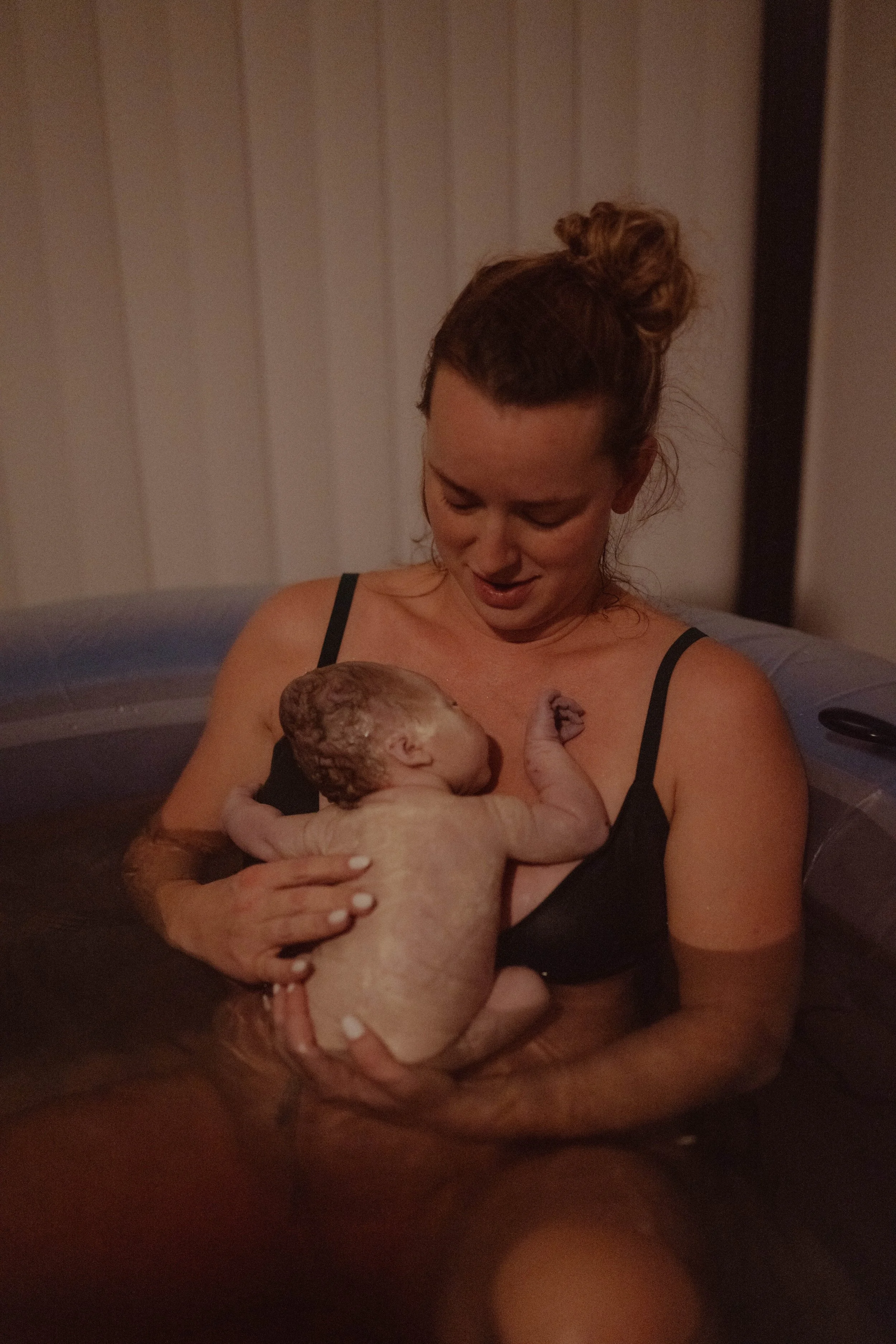 A woman in a black tank top is holding a newborn baby in a birthing tub. The woman is looking at the baby lovingly, and the baby is cuddled close to her chest.