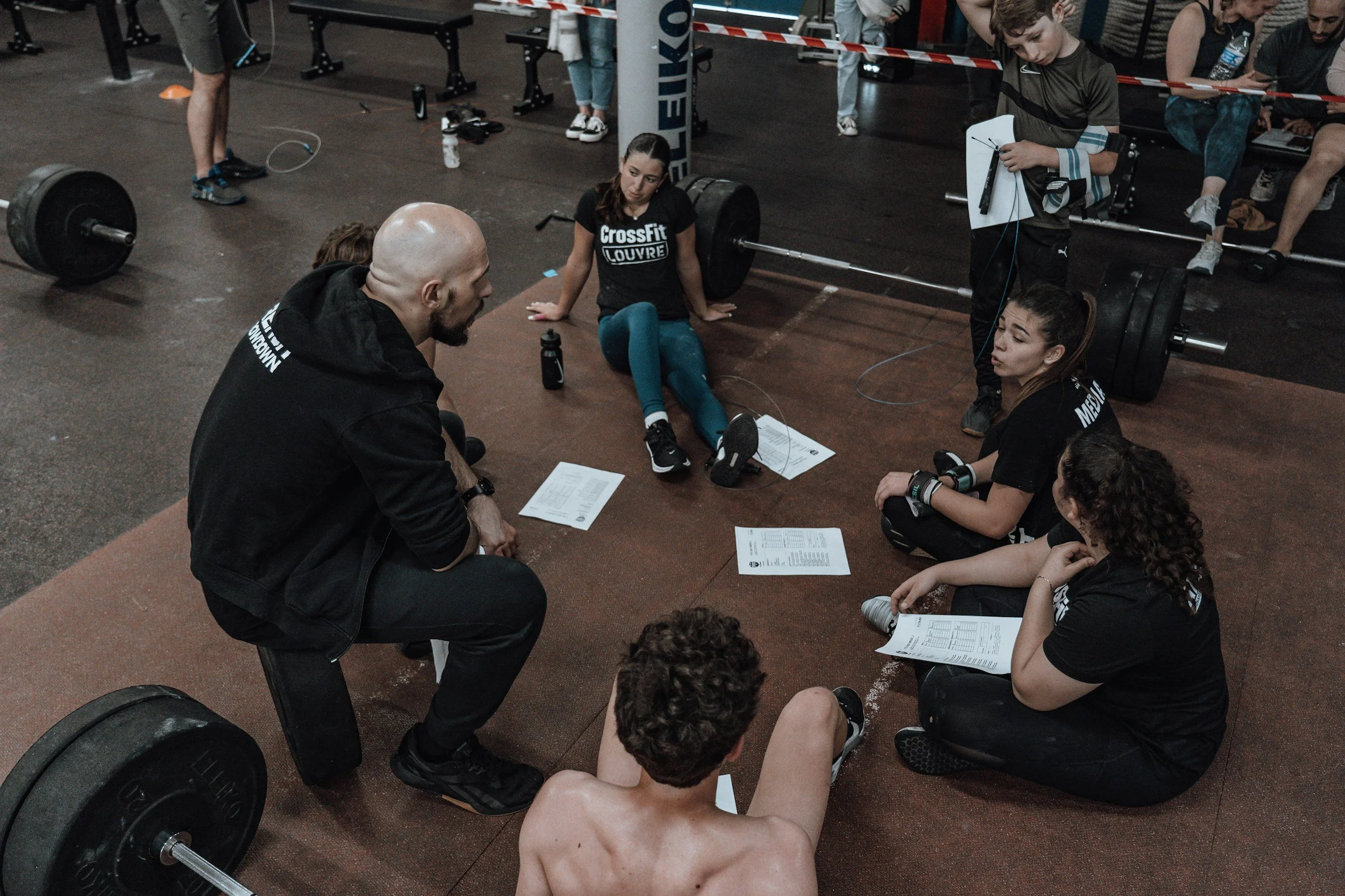 Group of athletes and coaches having a discussion on the gym floor during a CrossFit event, with some lifting weights and others seated with papers.