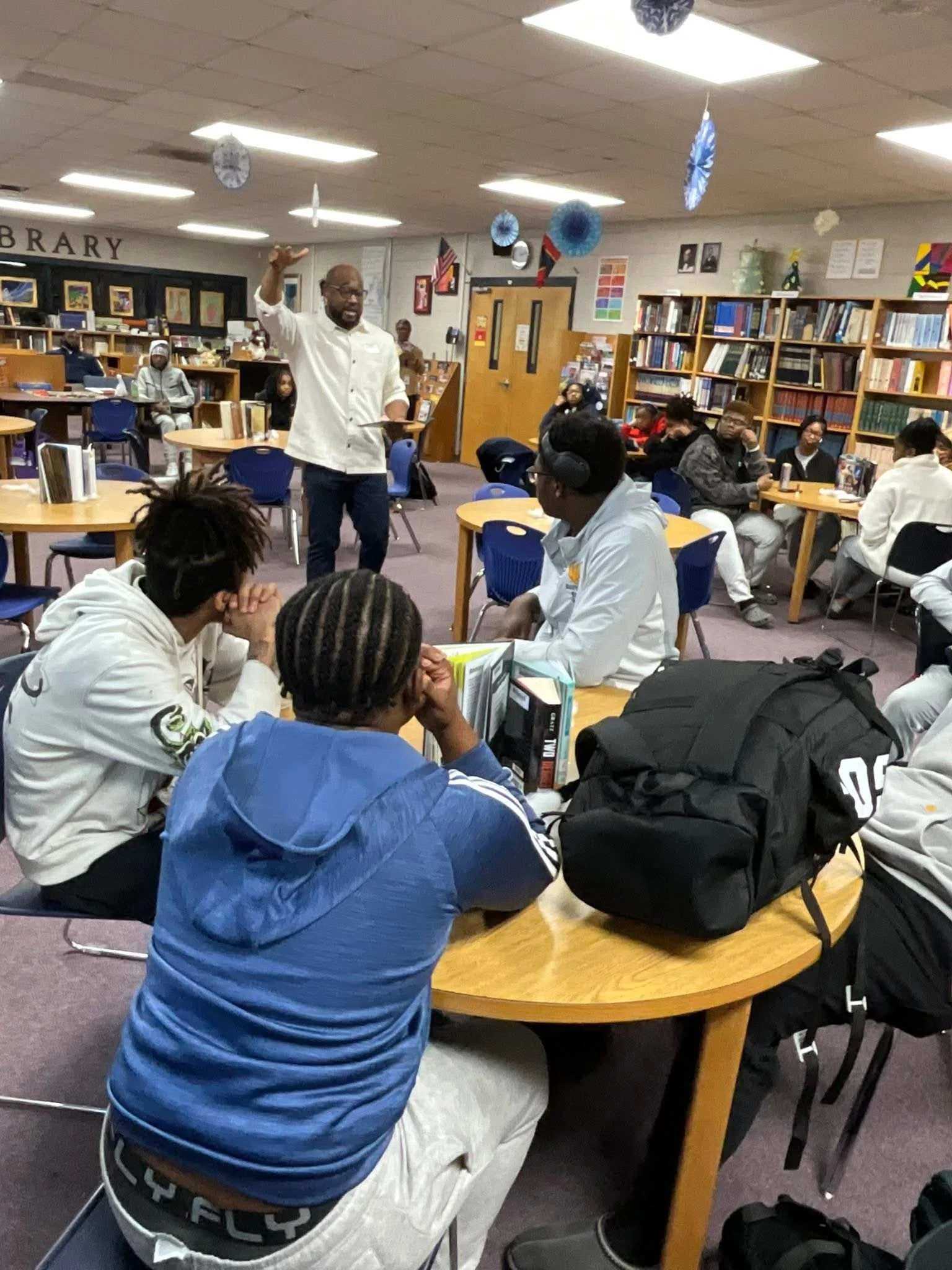 A man standing and speaking to a seated group in a library classroom. Several people are listening, some wearing headphones. Bookshelves and library decorations are visible in the background.