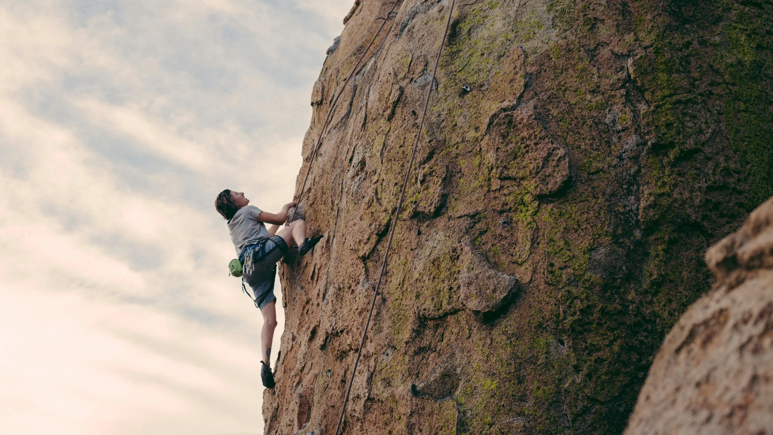 A person climbing an outdoor rock wall, wearing climbing gear, with an overcast sky in the background.