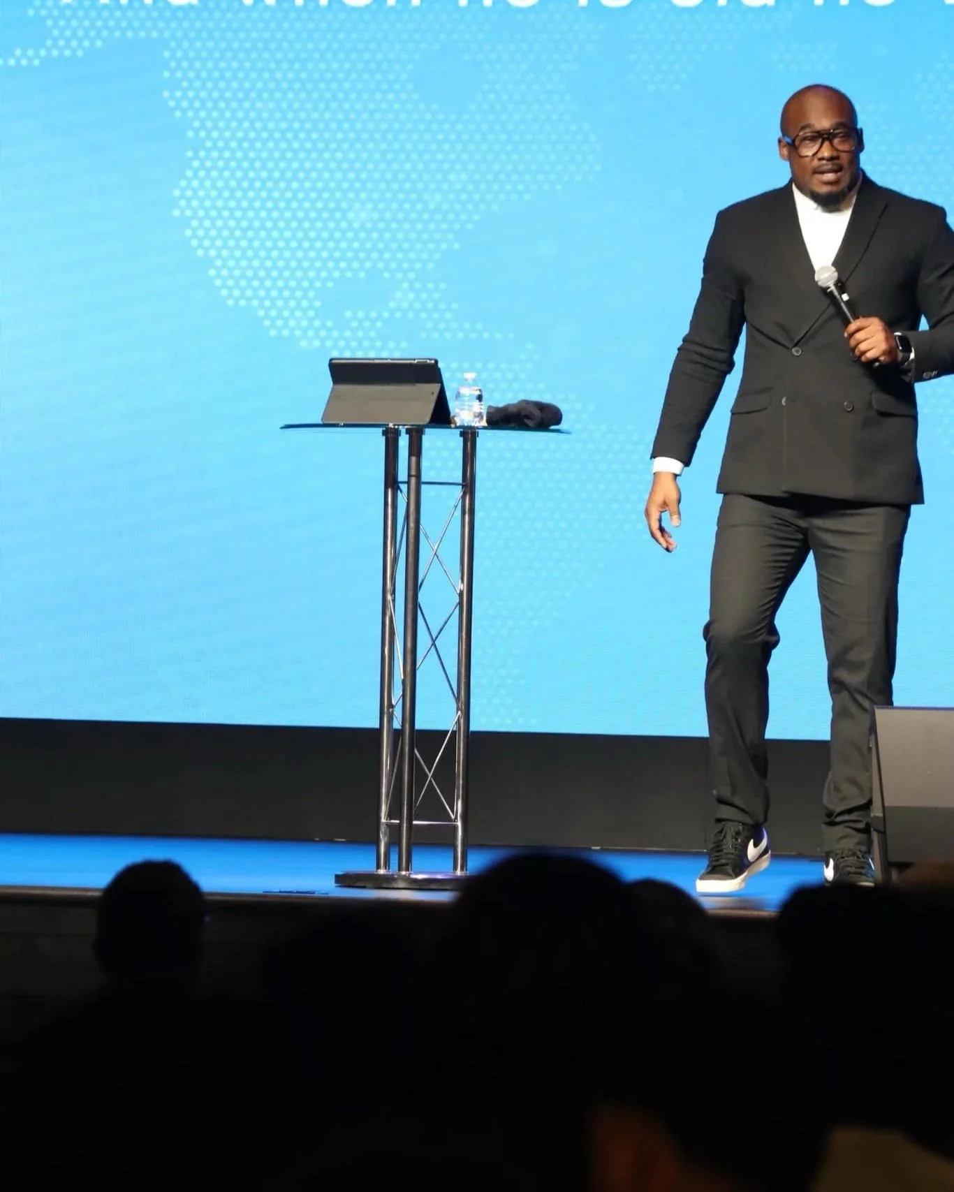 A man in a black suit holding a microphone on stage during a presentation, with a blue background and a podium with a water bottle and a tablet.