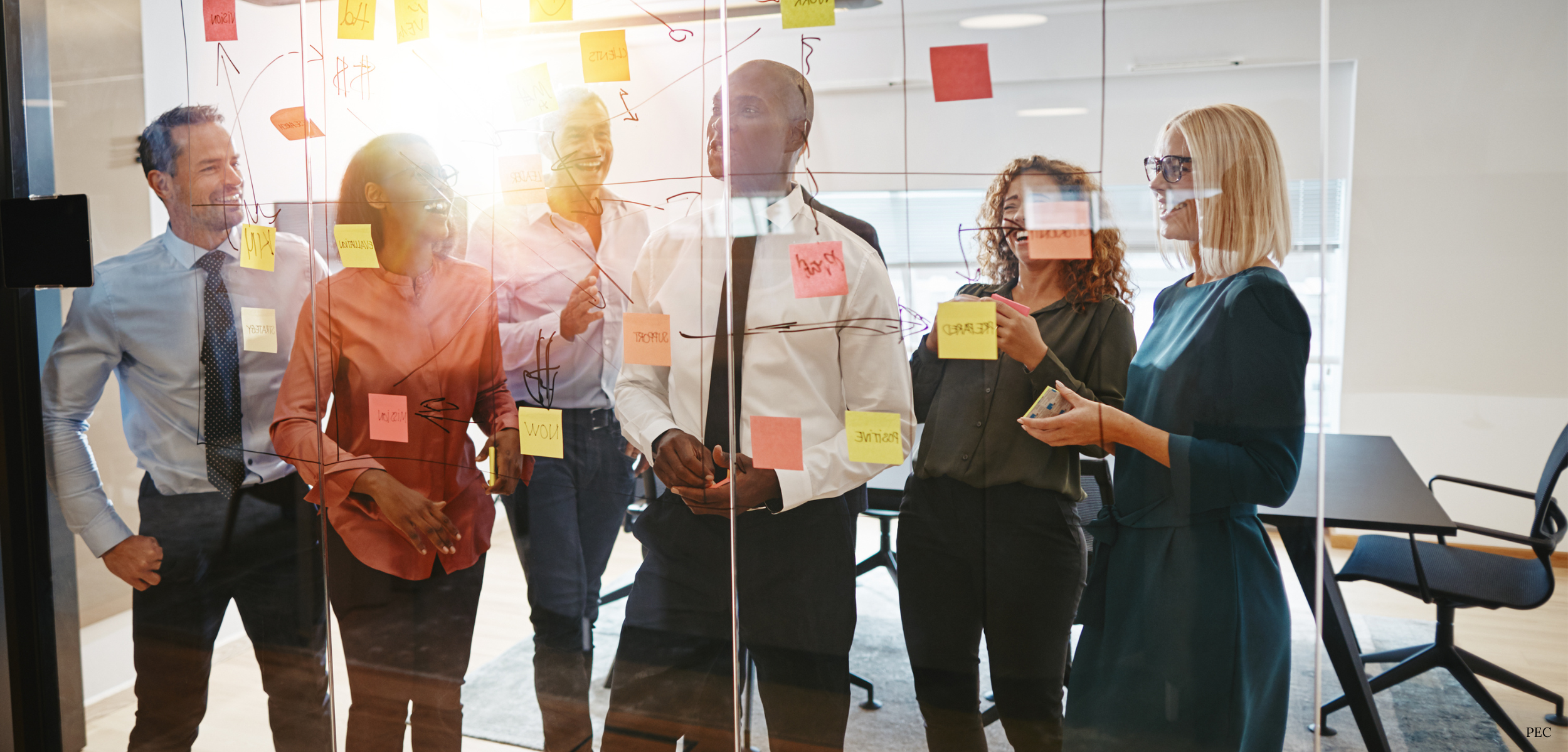A diverse group of six people in business attire standing behind a glass wall covered with colorful sticky notes and diagrams, engaging in a discussion and smiling in a modern office setting.