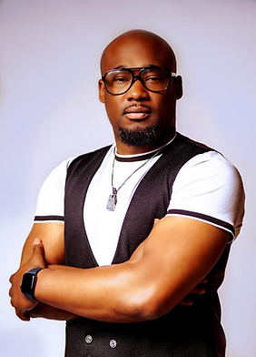 Portrait of a man with glasses and a beard, wearing a black vest over a white t-shirt, standing with arms crossed against a plain light background.