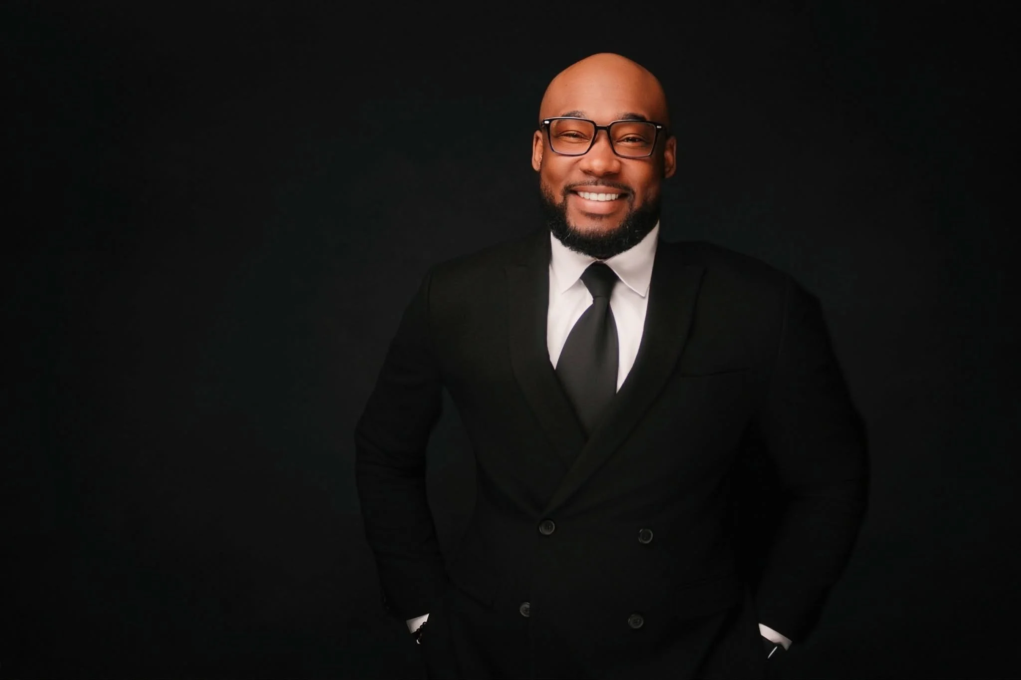 A man in a black suit holding a microphone on stage during a presentation, with a blue background and a podium with a water bottle and a tablet.