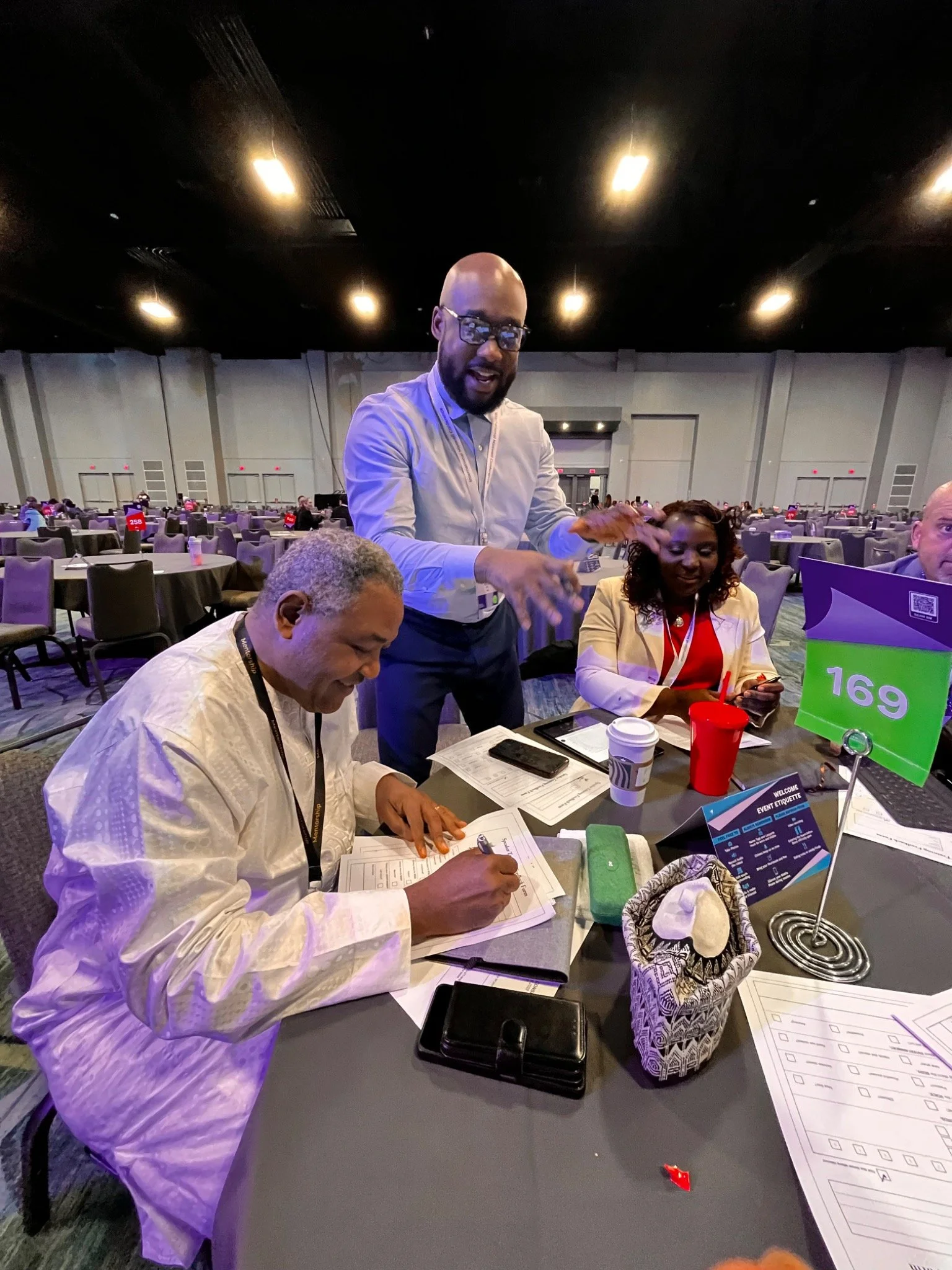Three people sitting at a round table in a large conference room, with one man standing and speaking, while the others are looking at papers or a phone. The table has a sign labeled '16g' and various documents, a coffee cup, and a small bag.
