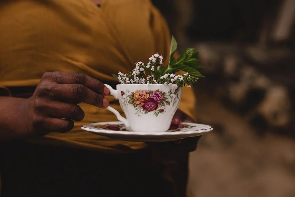 A person with dark skin wearing a yellow shirt holds a floral teacup with matching saucer, filled with small white flowers and green leaves.