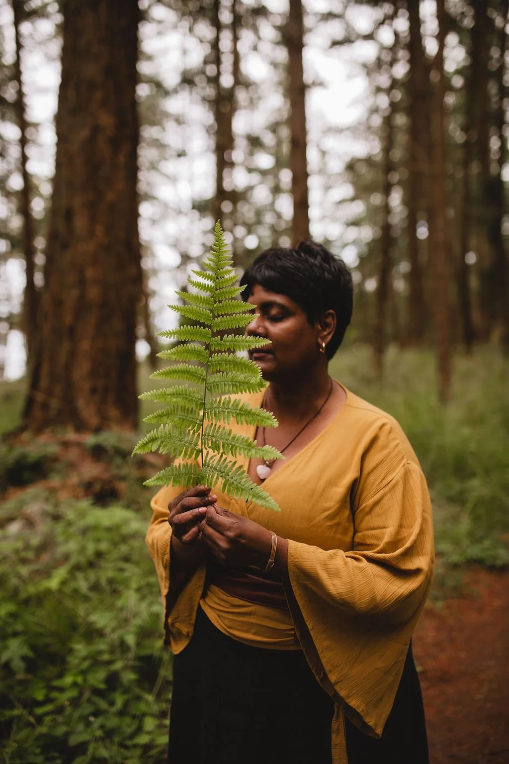A woman with short black hair and dark skin is holding a fern in a forest, with tall trees and greenery in the background.