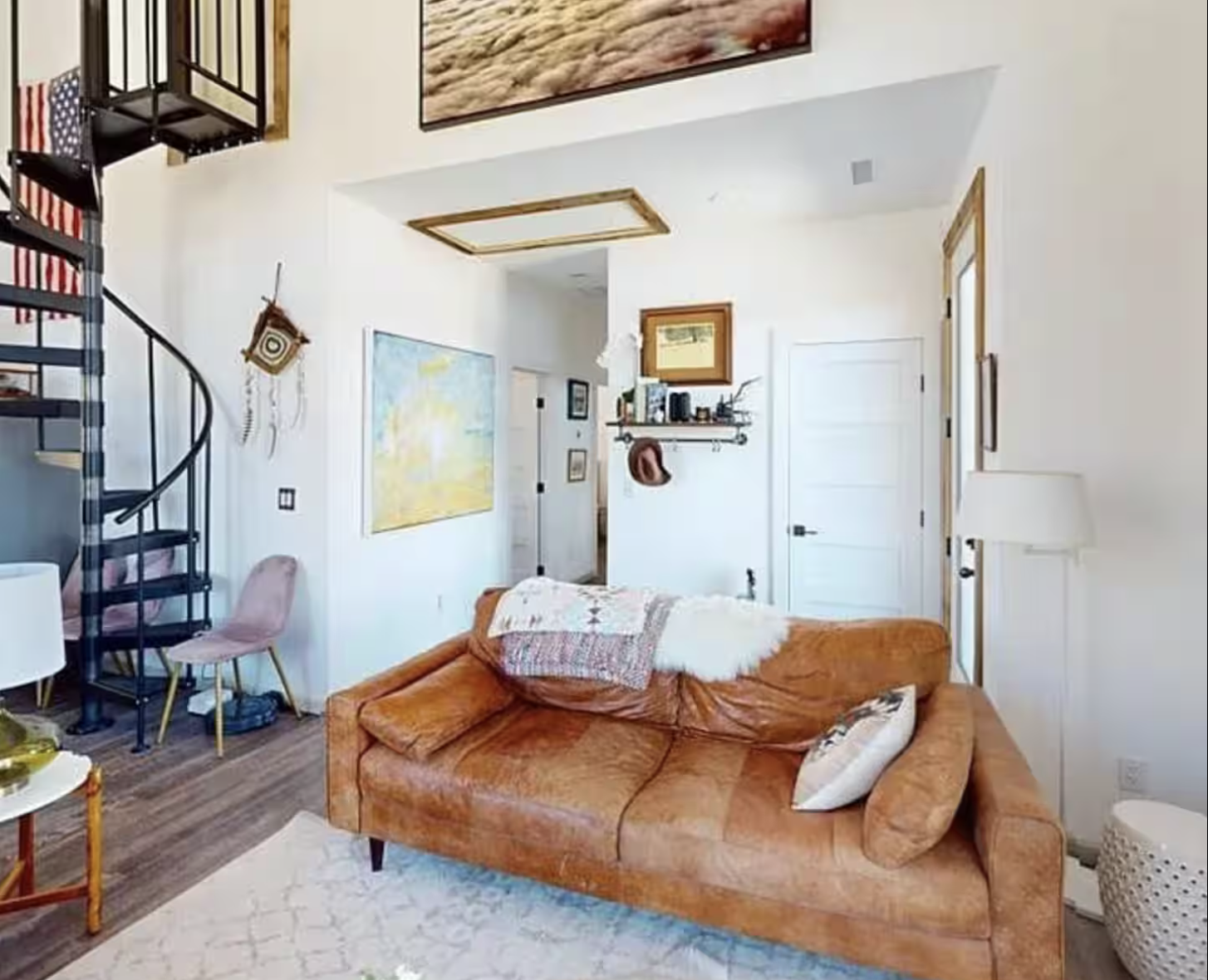 Living room with an orange sofa, a white lamp, a decorated pillow, and a blanket. A spiral staircase, wall art, and a small pink chair are visible. The room has wood flooring, white walls, and various framed pictures and decorations.