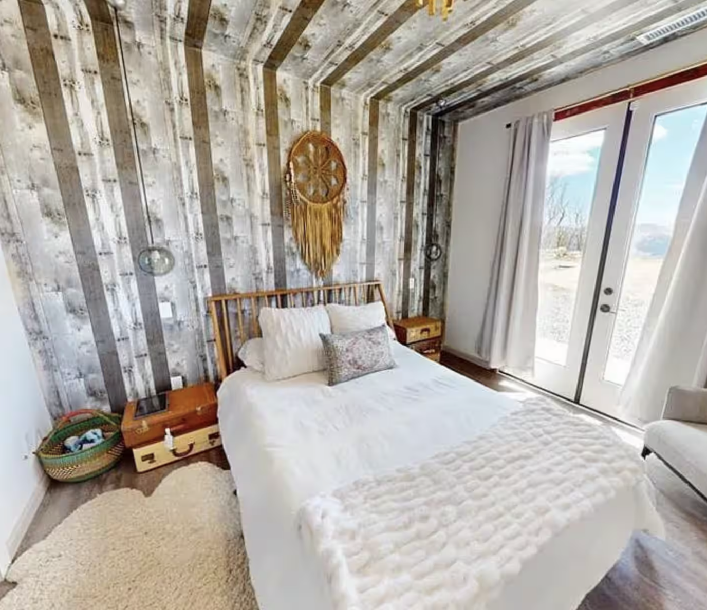 A bedroom with a wooden bed frame, white bedding, pillows, wall hanging, nightstands, and a sliding glass door with white curtains.