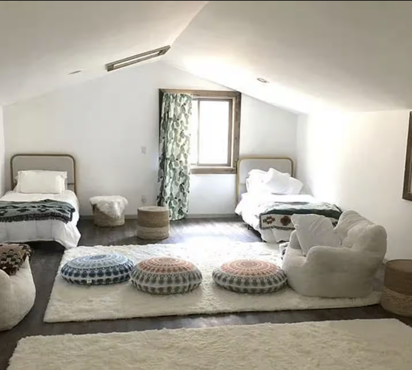 Cozy attic bedroom with two beds, a white plush armchair, colorful round cushions, and a window with floral curtains.