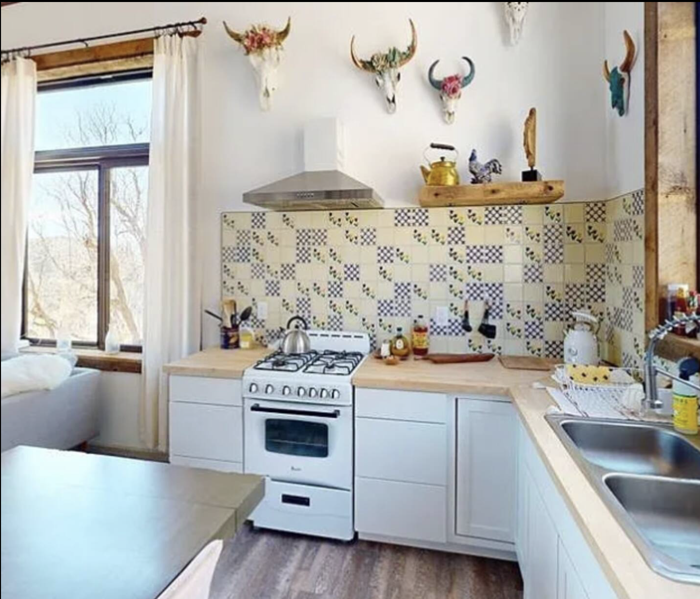 Kitchen with white cabinets, a stove, a sink, and decor on the wall including animal skulls with floral arrangements, along with a window with white curtains and a view of trees outside.