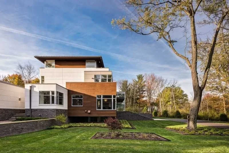 Modern multi-story house with mixed siding of white and wood, large windows, and landscaped front yard with trees and grass.