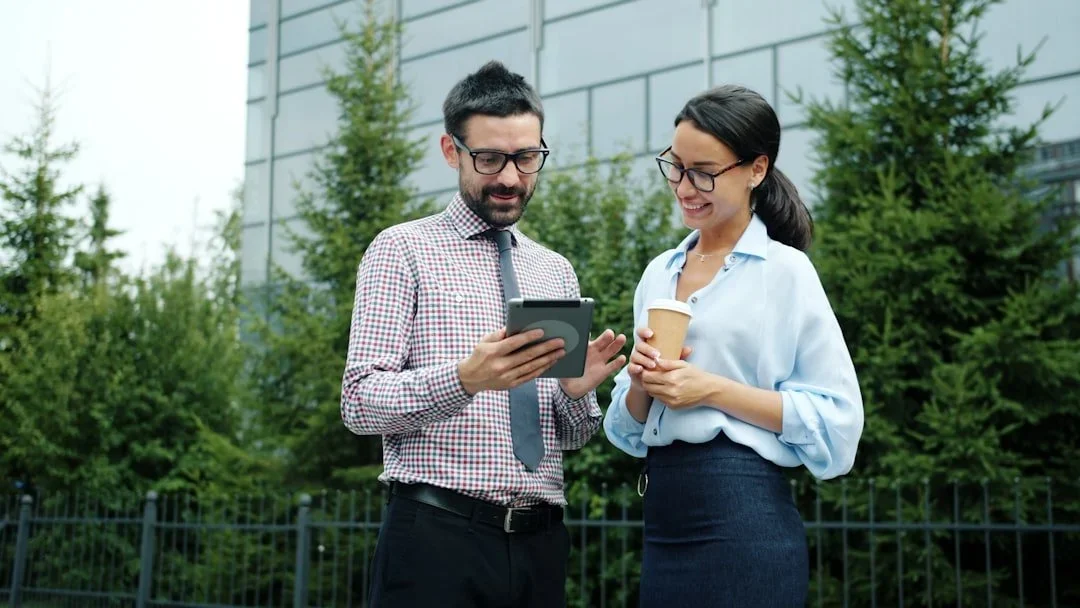 A man and woman in business attire outside, looking at a smartphone together. The woman is holding a takeaway coffee cup and smiling. They are standing in front of a modern building with trees around.