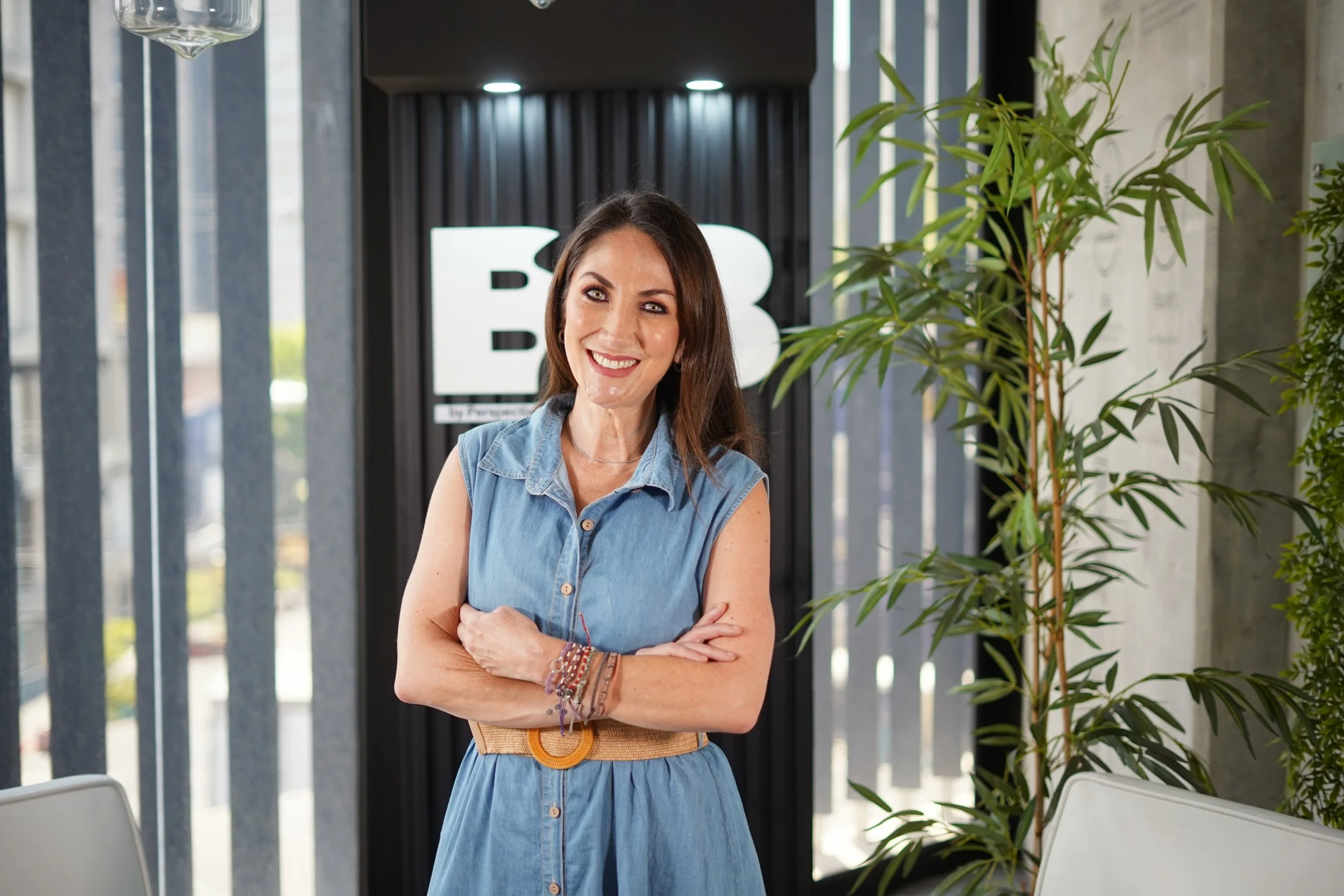 Una mujer sonriendo con los brazos cruzados, lleva una blusa de mezclilla y un cinturón beige, con plantas y un fondo de oficina moderna con letras 'B' y 'U'.