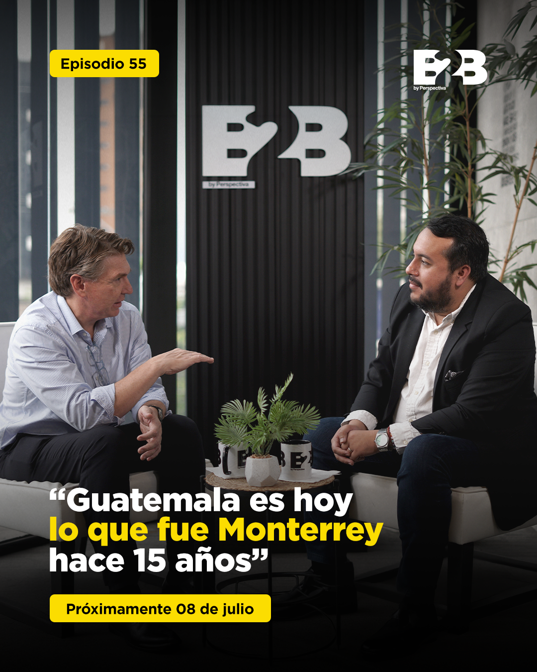 Dos hombres sentados en una mesa de oficina en una conversación, con un fondo con signos de BB y plantas de interior en un ambiente moderno.