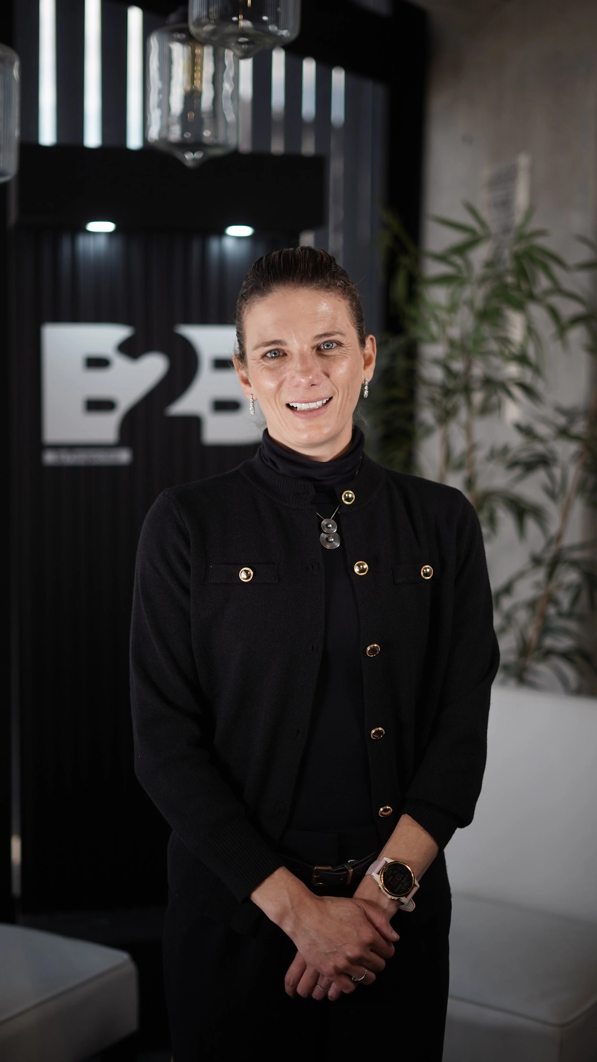 Retrato de una mujer sonriendo, de cabello oscuro y coleta, vestida con un suéter negro, en un entorno de oficina con plantas y logotipo de fondos.