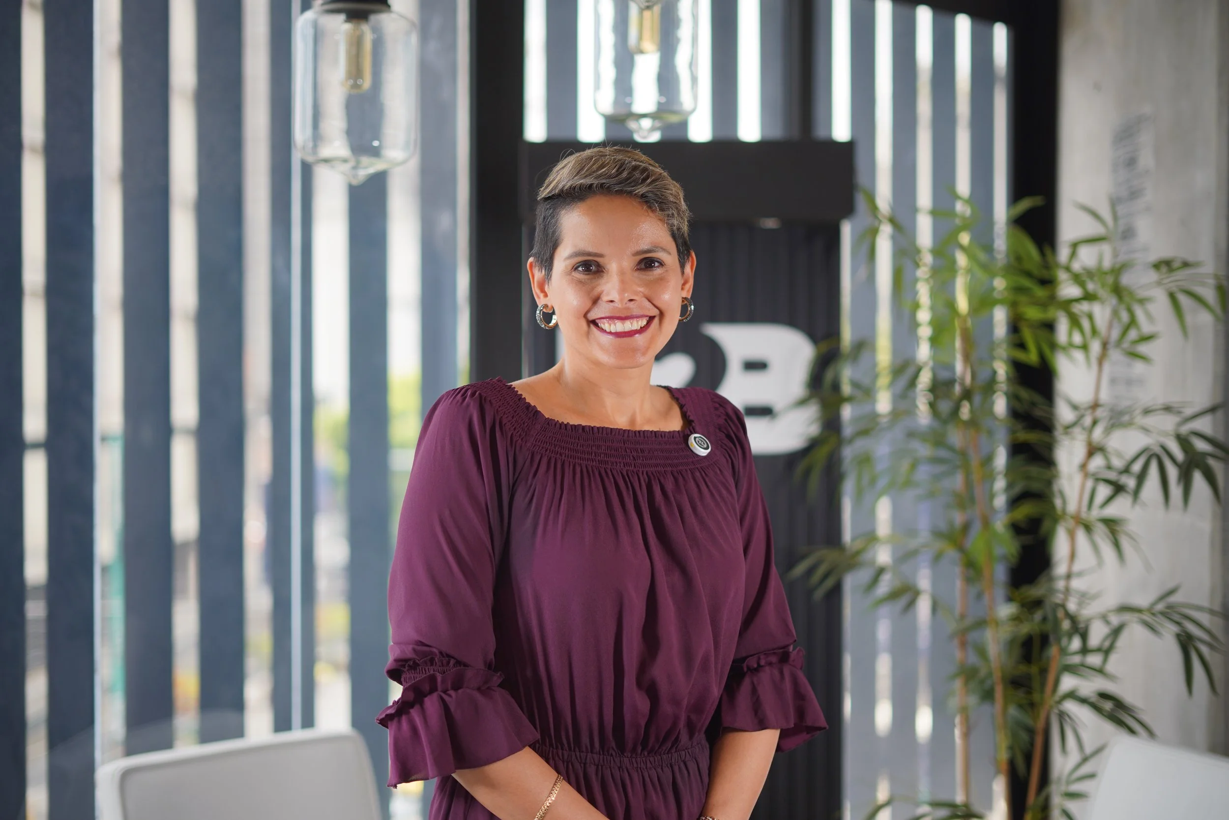 Mujer sonriendo, con vestido vino y pendientes, en un ambiente moderno con plantas y decoraciones.