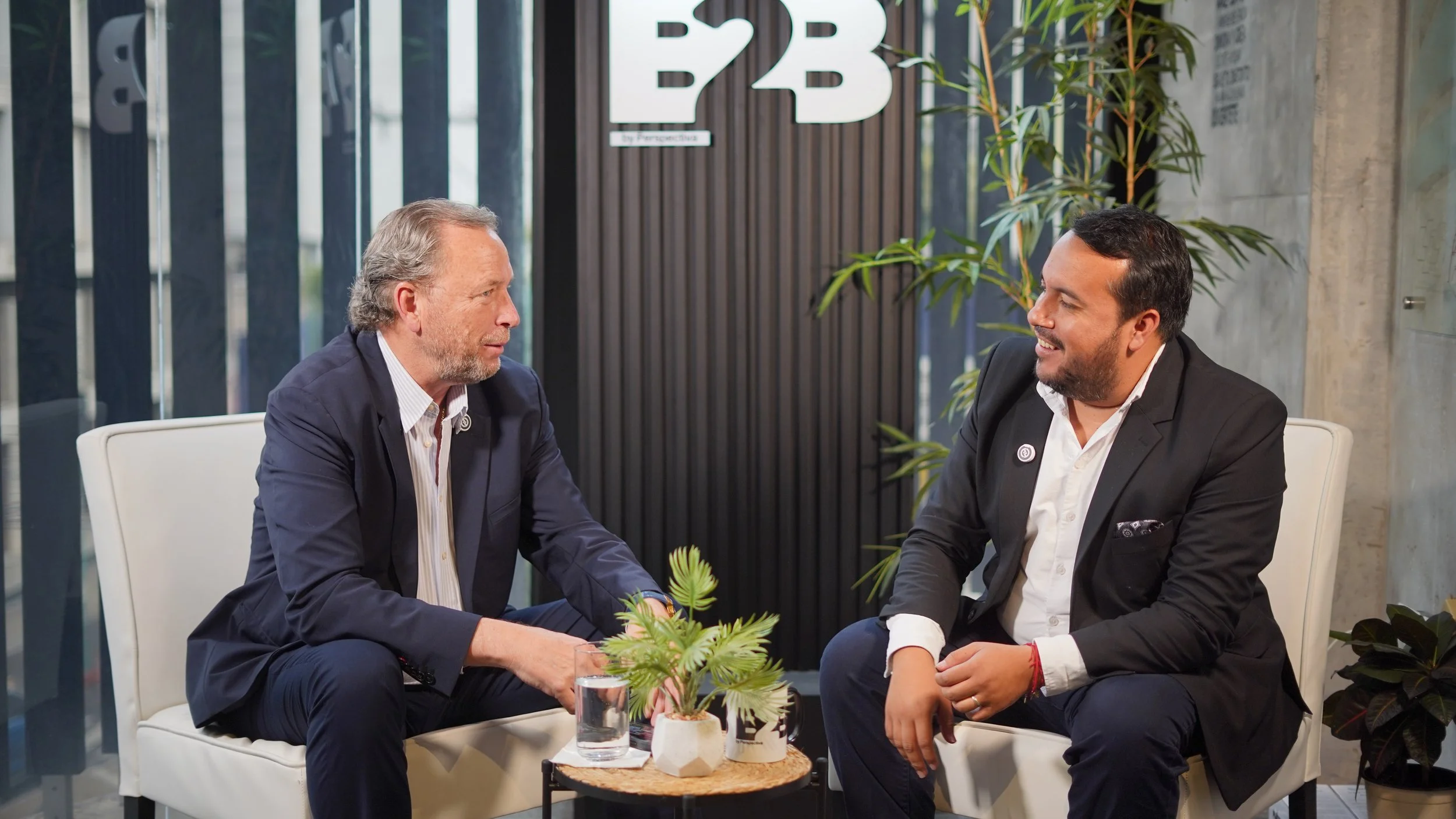 Dos hombres de negocios conversando en una sala moderna con plantas y logotipo de B2B en la pared.
