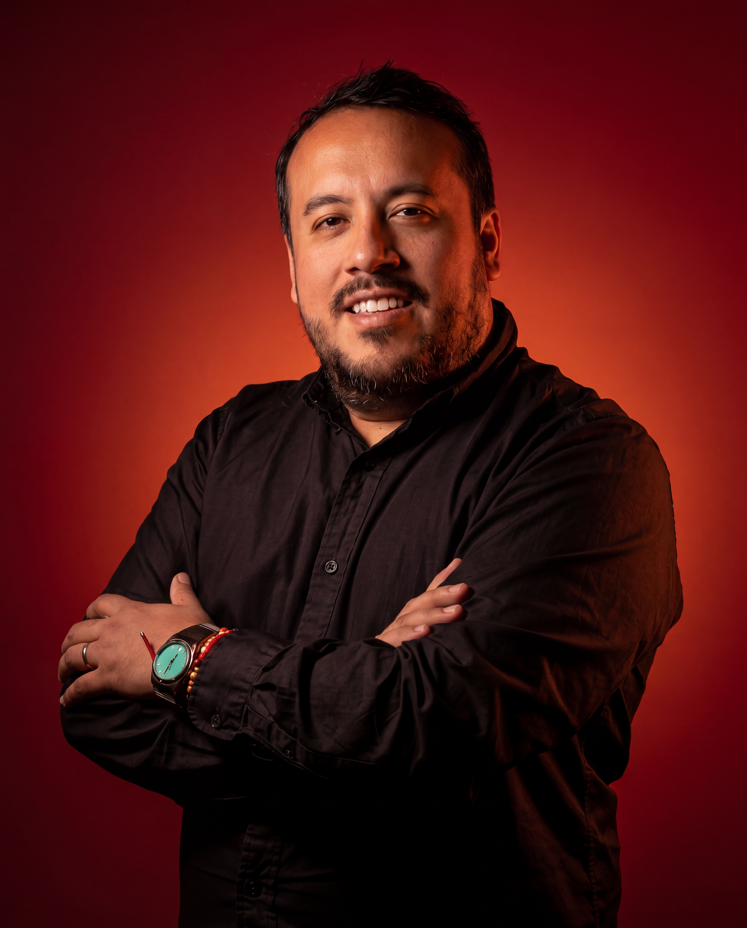 Joven hombre con barba y sonrisa, cruzado de brazos, fondo rojo oscuro.
