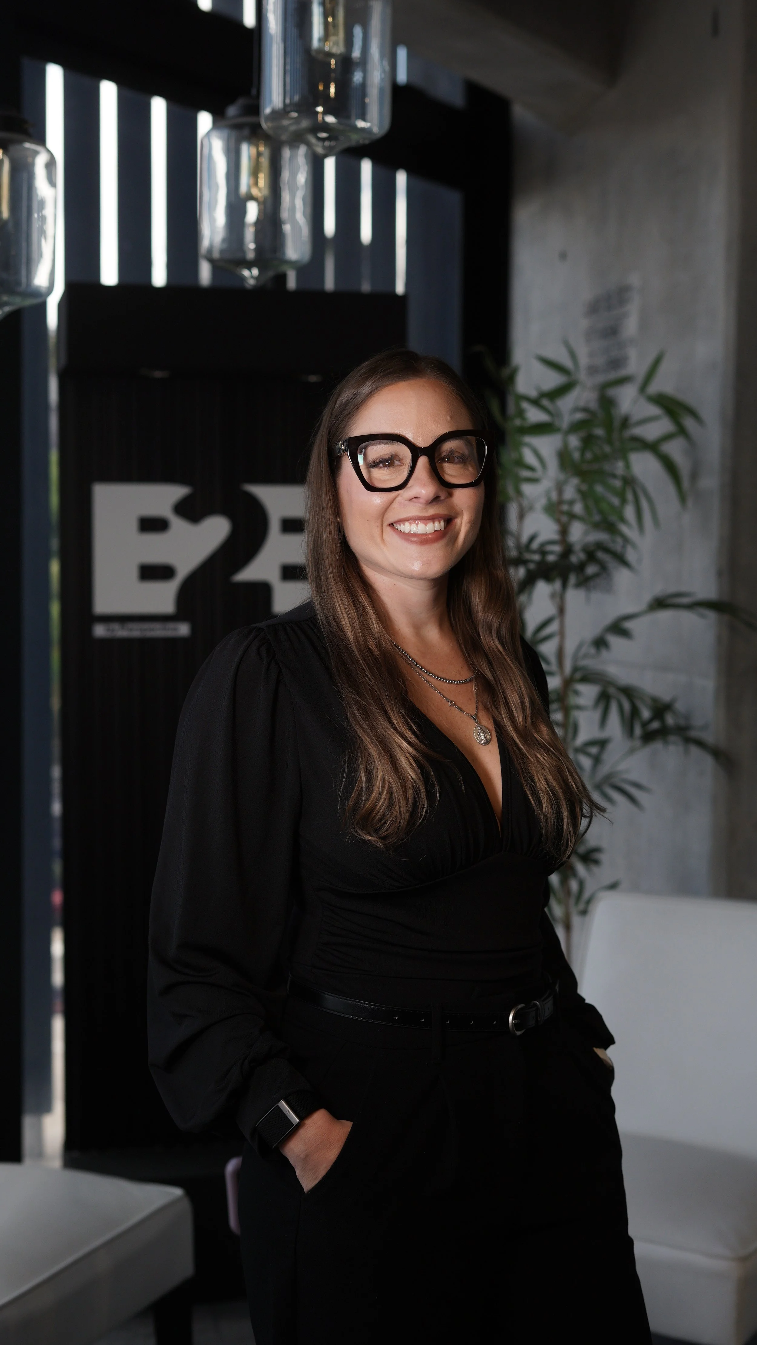 Mujer sonriendo, usando gafas grandes y ropa negra, posando en un espacio moderno con plantas y decoración.