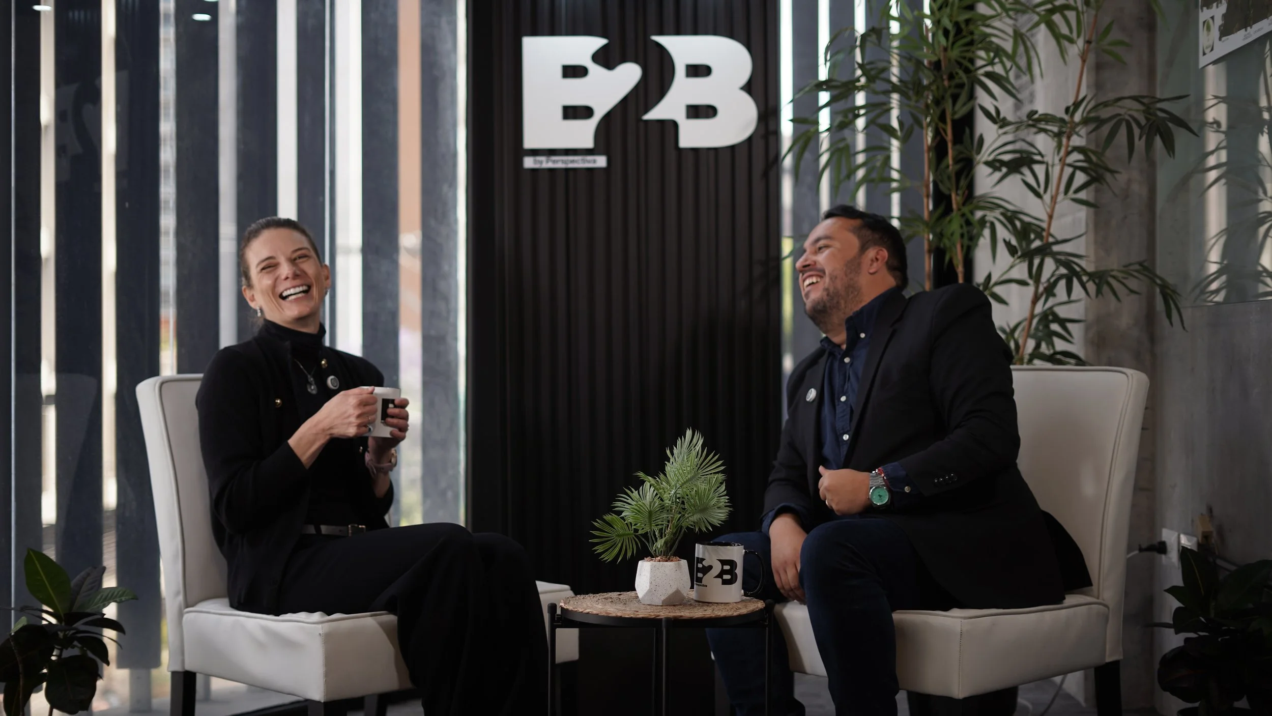 Dos personas, un hombre y una mujer, sentadas en una sala moderna, riendo y conversando, con tazas en la mano, en un ambiente de oficina o lounge, con plantas y un cartel de logo B2B en el fondo.