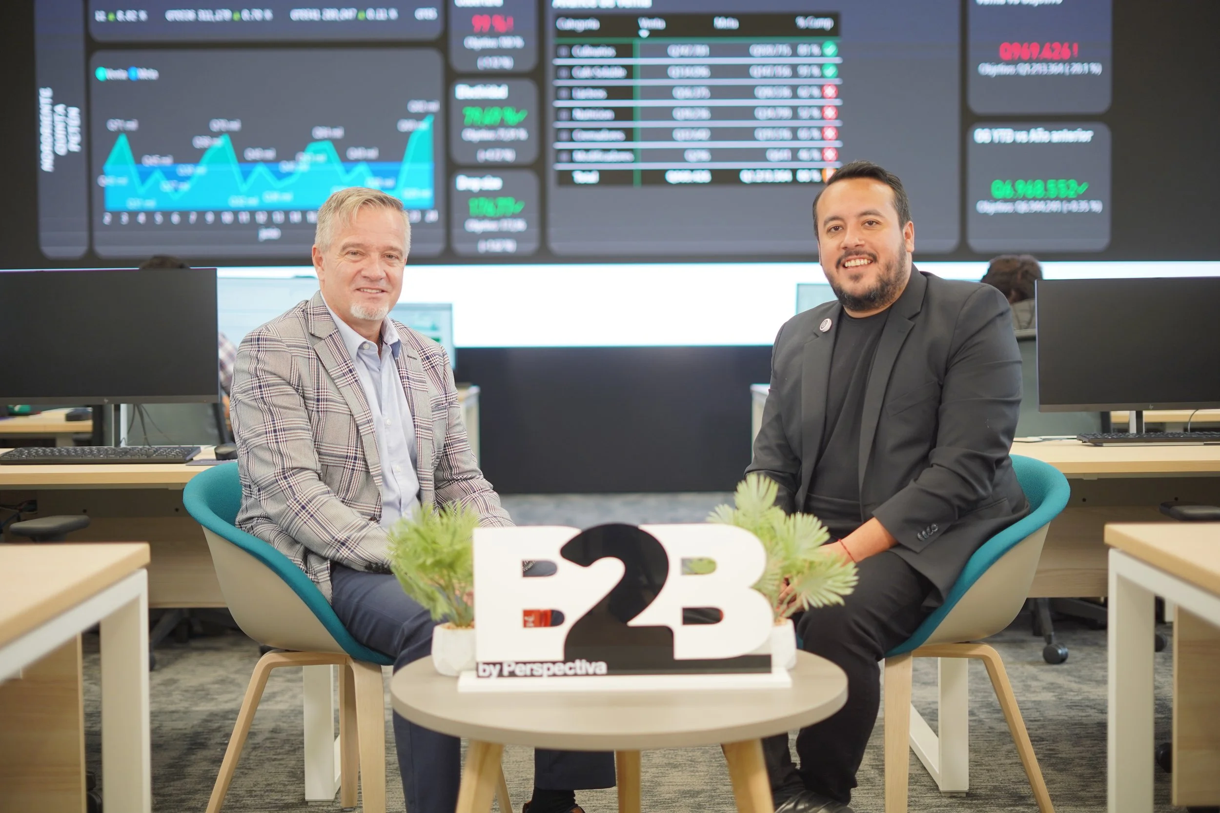 Dos hombres sentados en una mesa en un ambiente de oficina, con pantallas de datos financieros en el fondo.