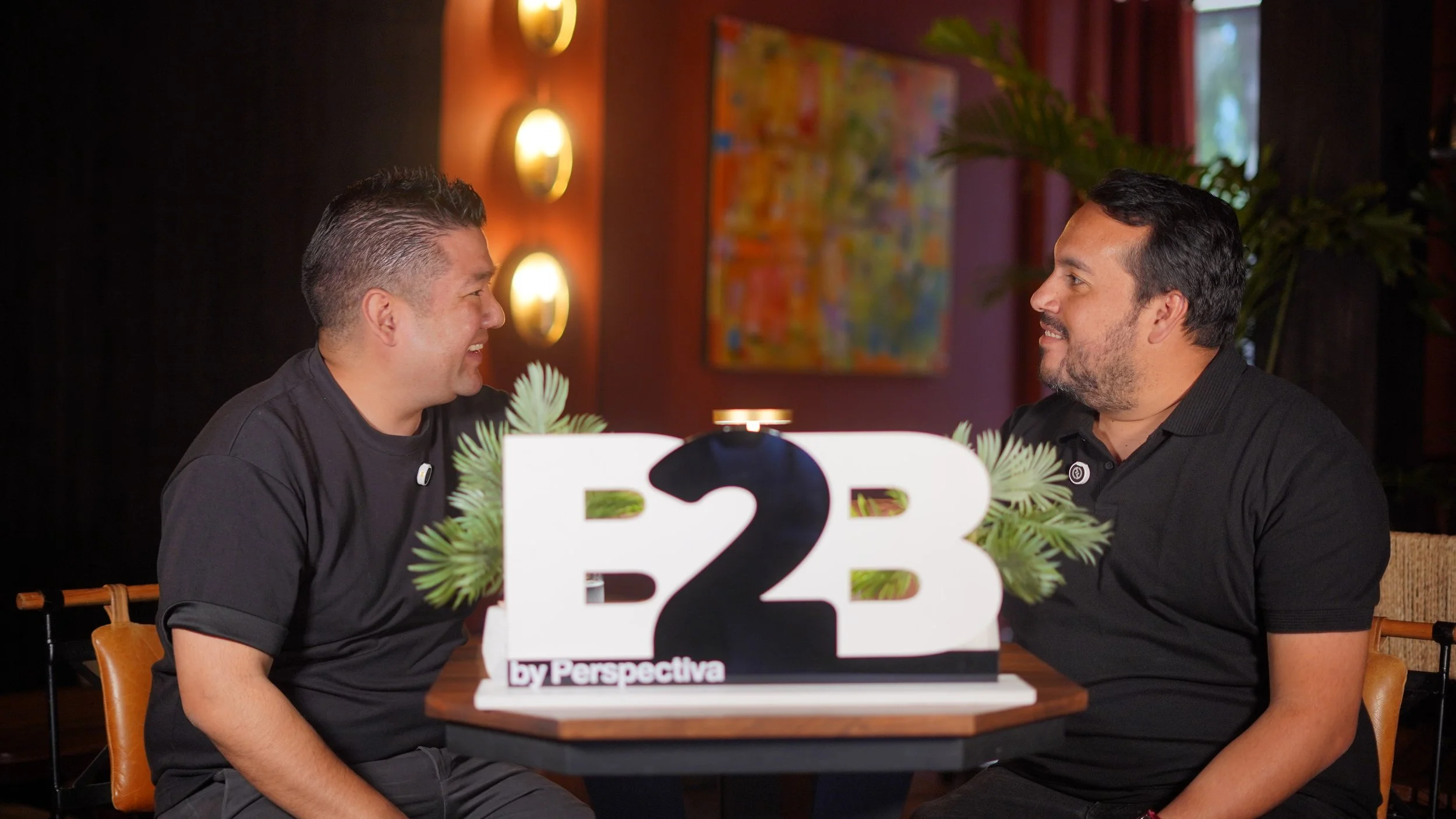Dos hombres sentados en una mesa de restaurante, sonriendo y mirándose, con cartel que dice 'B2B' en la mesa.