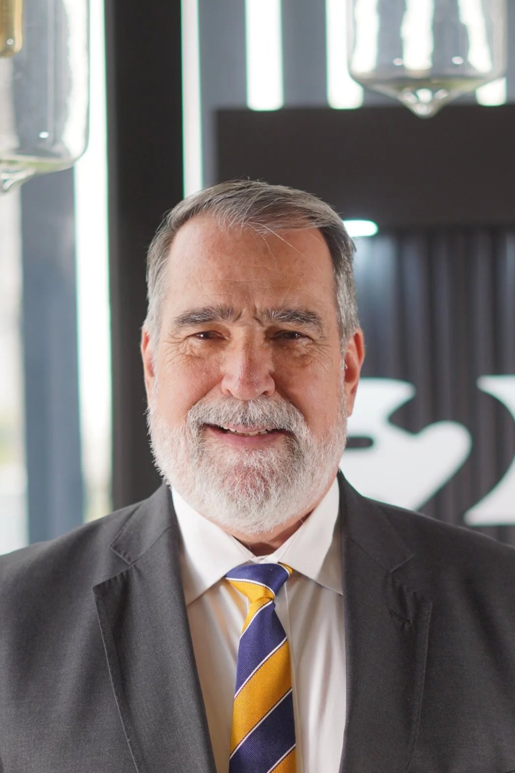 Hombre mayor con barba y bigote, vestido con traje y corbata de rayas azules, amarillas y blancas, sonriendo en un entorno moderno.
