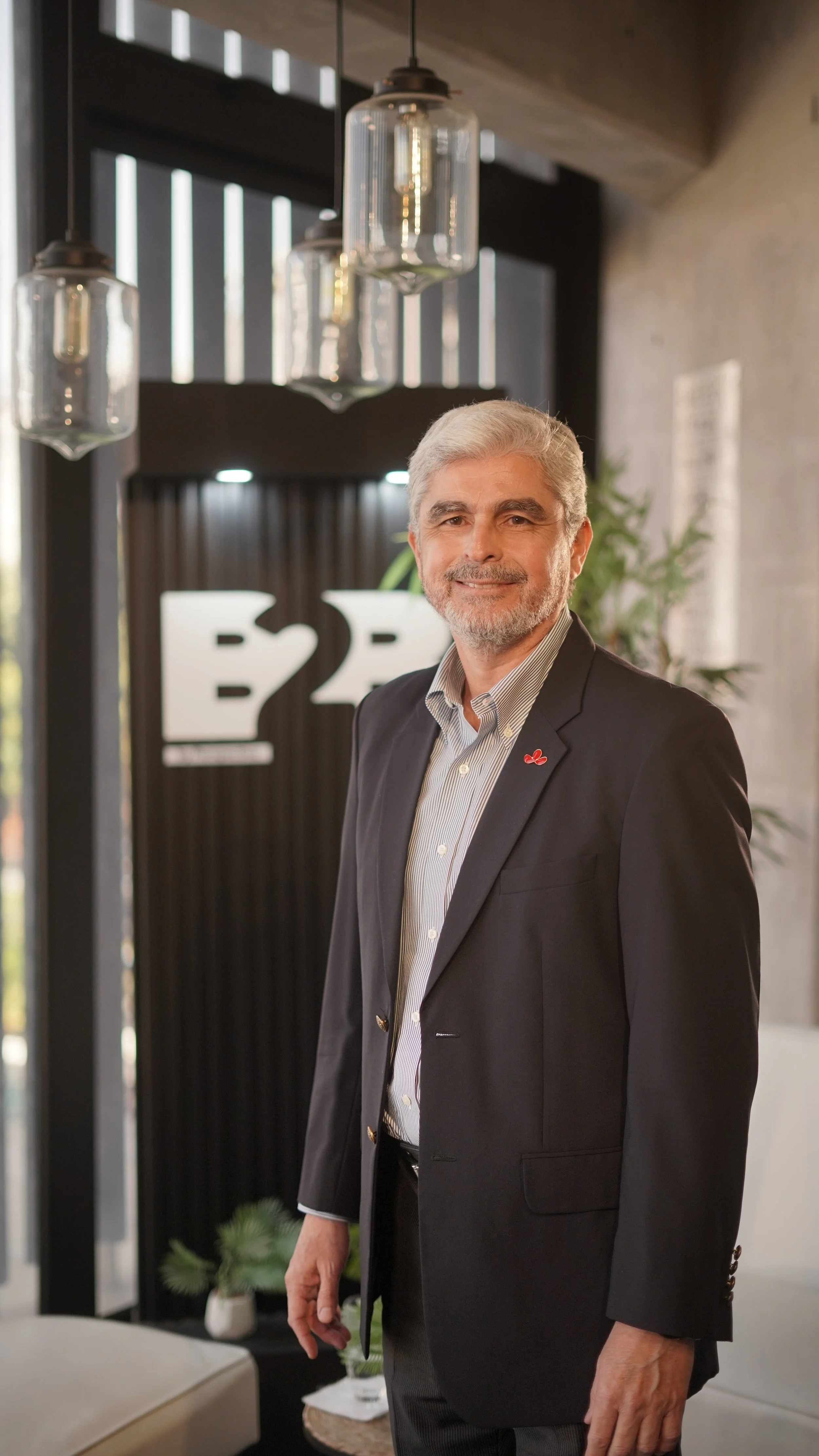 Hombre de avanzada edad con cabello gris y barba, vestido con traje negro y camisa blanca, posando en un ambiente interior moderno con lámparas colgantes y una señal que dice 'E2F' en el fondo.