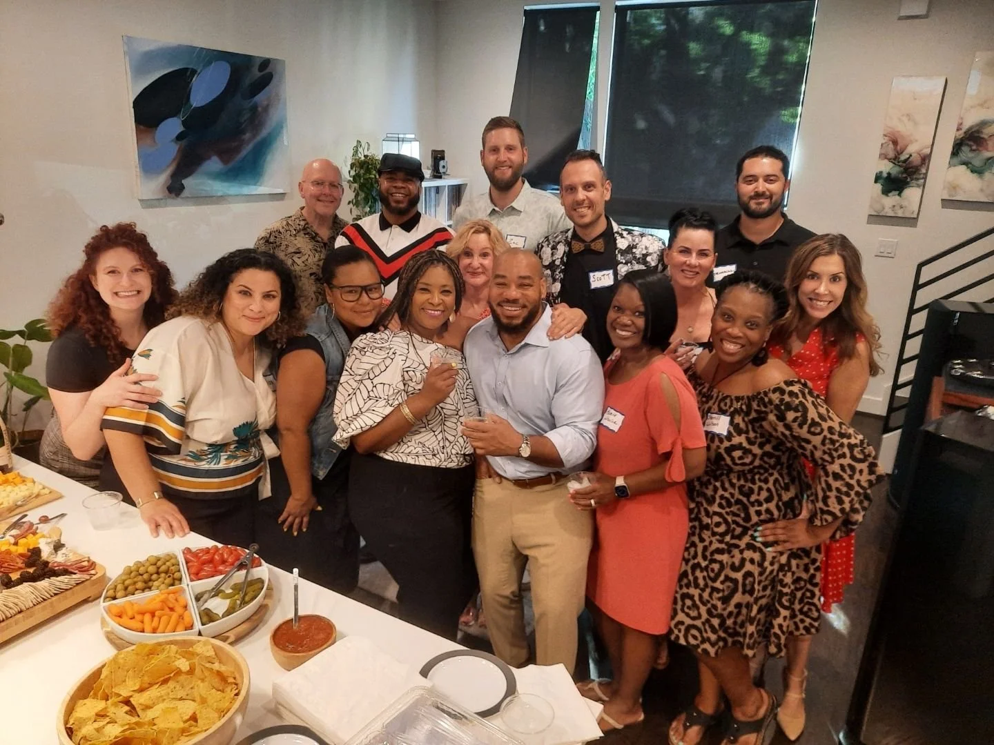 A group of 15 people gathered at a party, smiling and posing for a photo. There are platters of food on the table in front of them.