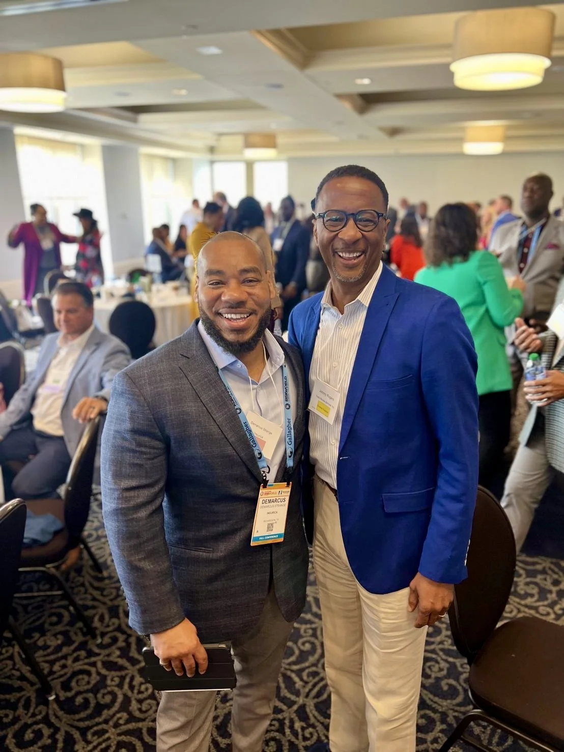Two men in suits smiling and standing close together at a conference or event, with other attendees in the background.