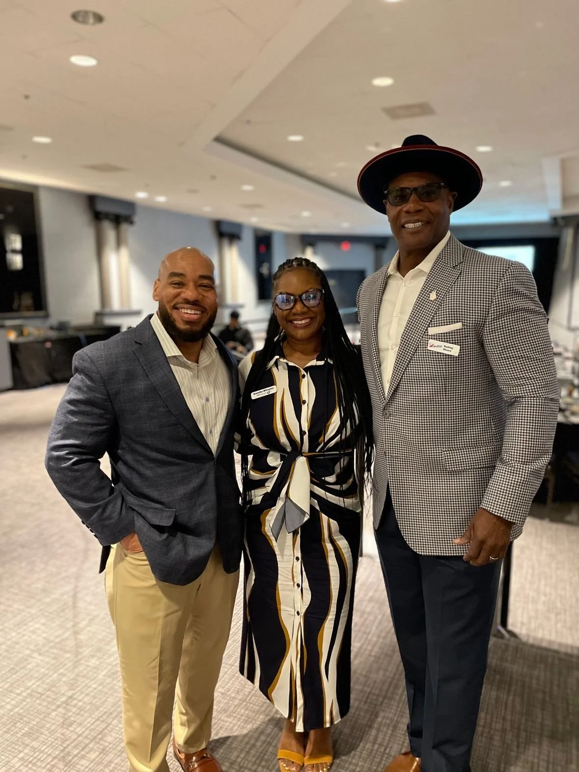 Three people dressed in formal attire standing together and smiling at the camera in an event space or restaurant. The man on the left is wearing a gray blazer, light-colored pants, and a striped shirt. The woman in the middle is wearing a black, white, and gold striped dress with glasses. The man on the right is taller, wearing a checkered blazer, white shirt, dark pants, glasses, and a wide-brimmed hat.