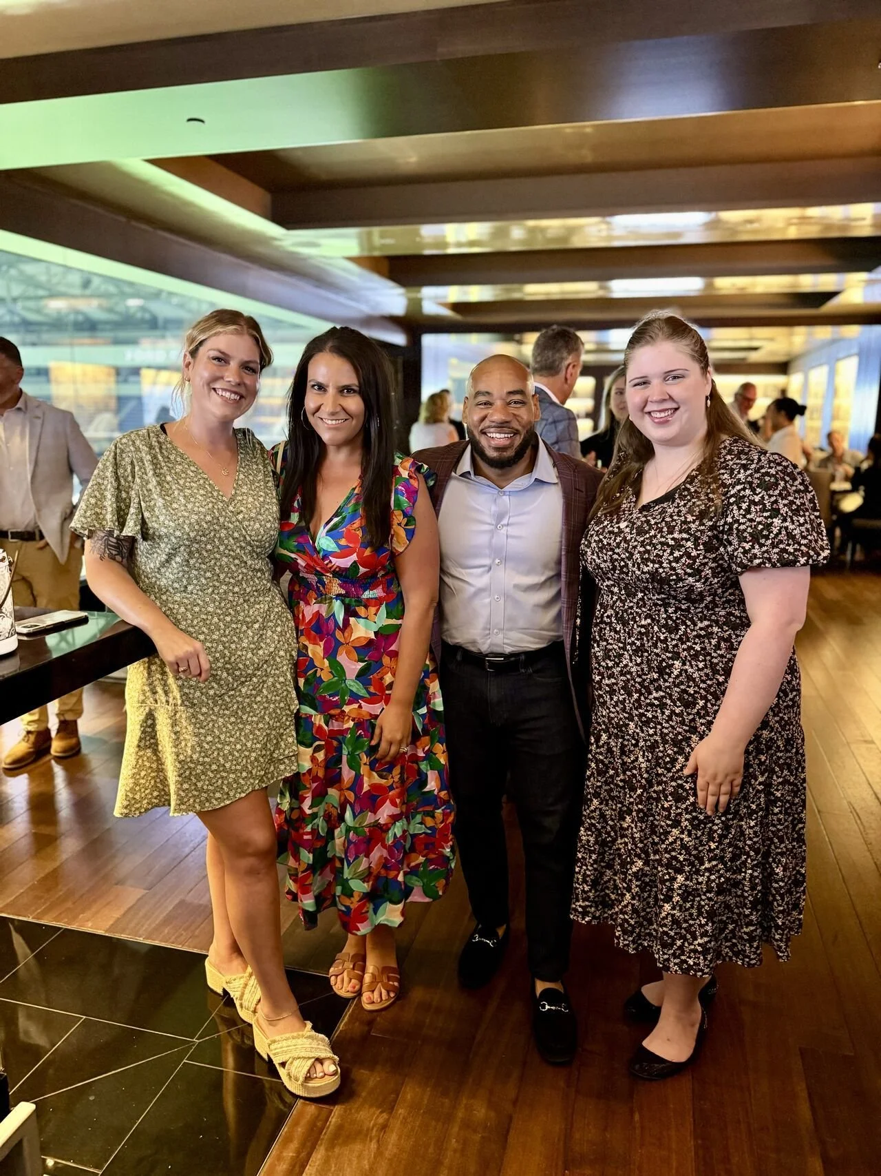 Four people, three women and one man, standing close together and smiling inside a modern restaurant or event space. The women are wearing floral dresses, and the man is in a suit with an open-collar shirt.