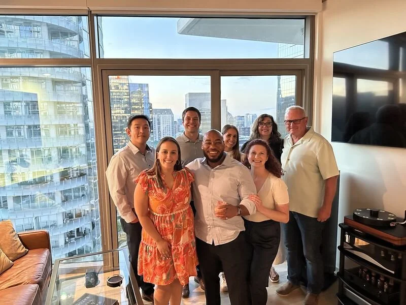 Group of eight diverse people posing together in a modern apartment with city view, smiling, one holding a drink.