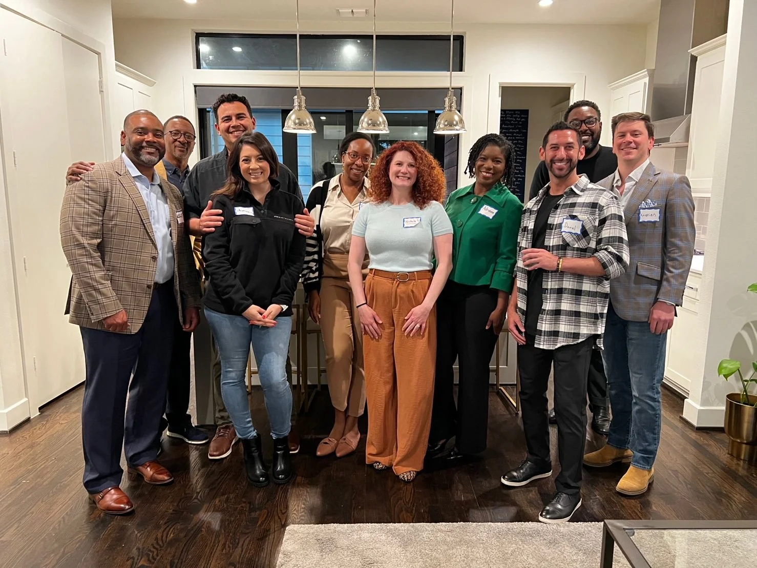A group of twelve diverse adults standing together in a kitchen, smiling at the camera during a social gathering or event.