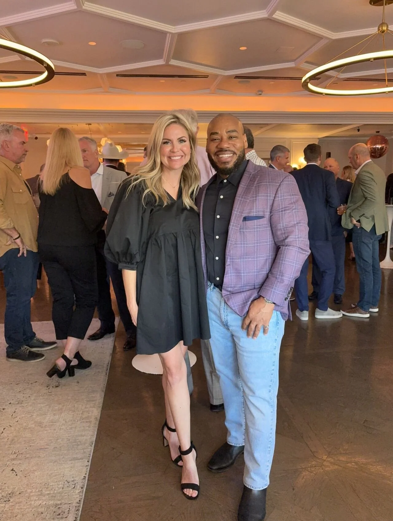 A woman in a black dress and high heels stands next to a man in a purple checkered blazer, black shirt, and light-wash jeans at a social event or party with other guests in the background.