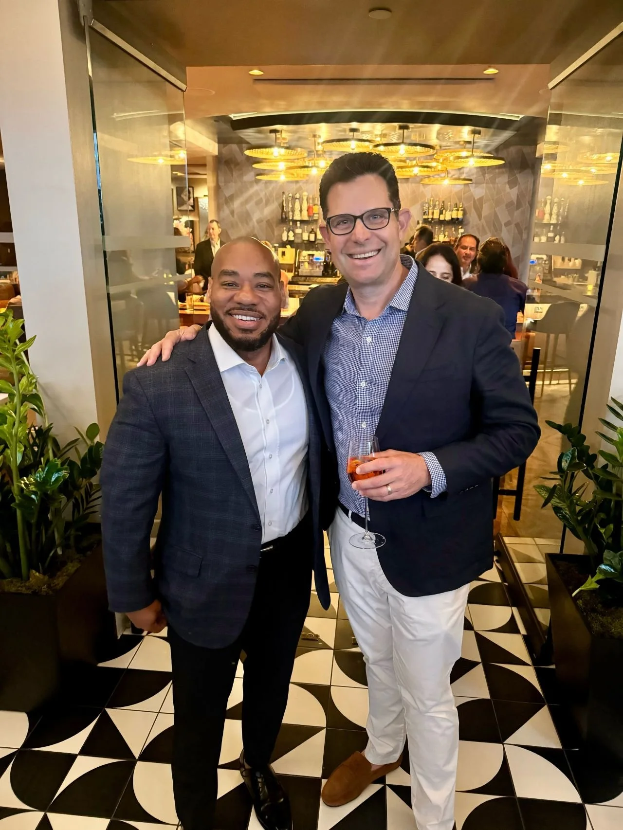 Two men in formal attire smiling at a restaurant or bar with a background of shelves filled with bottles, with other people visible in the background.